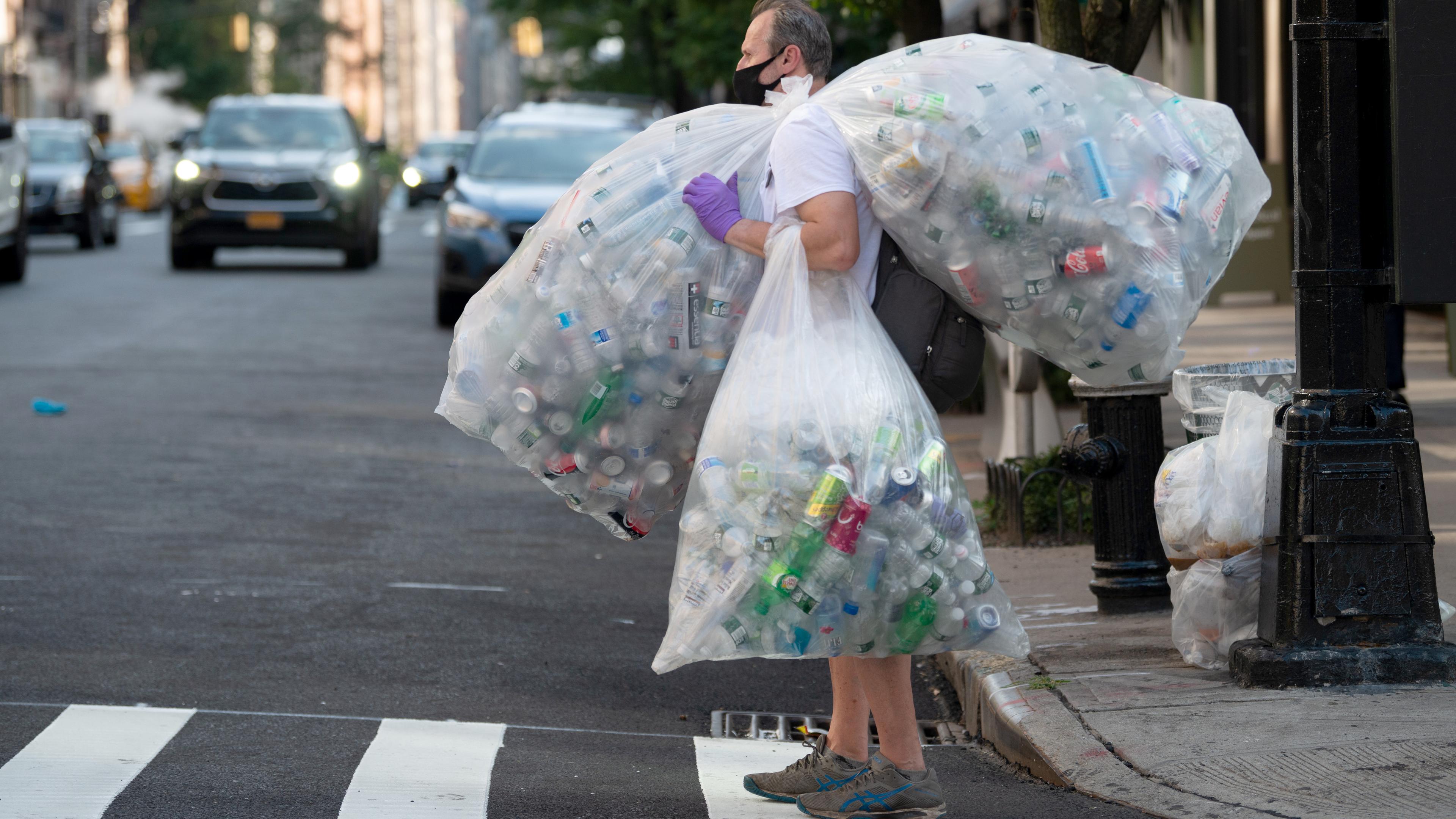 Photo of a person with a face mask carrying large bags of plastic bottles on a city street with cars in the background.