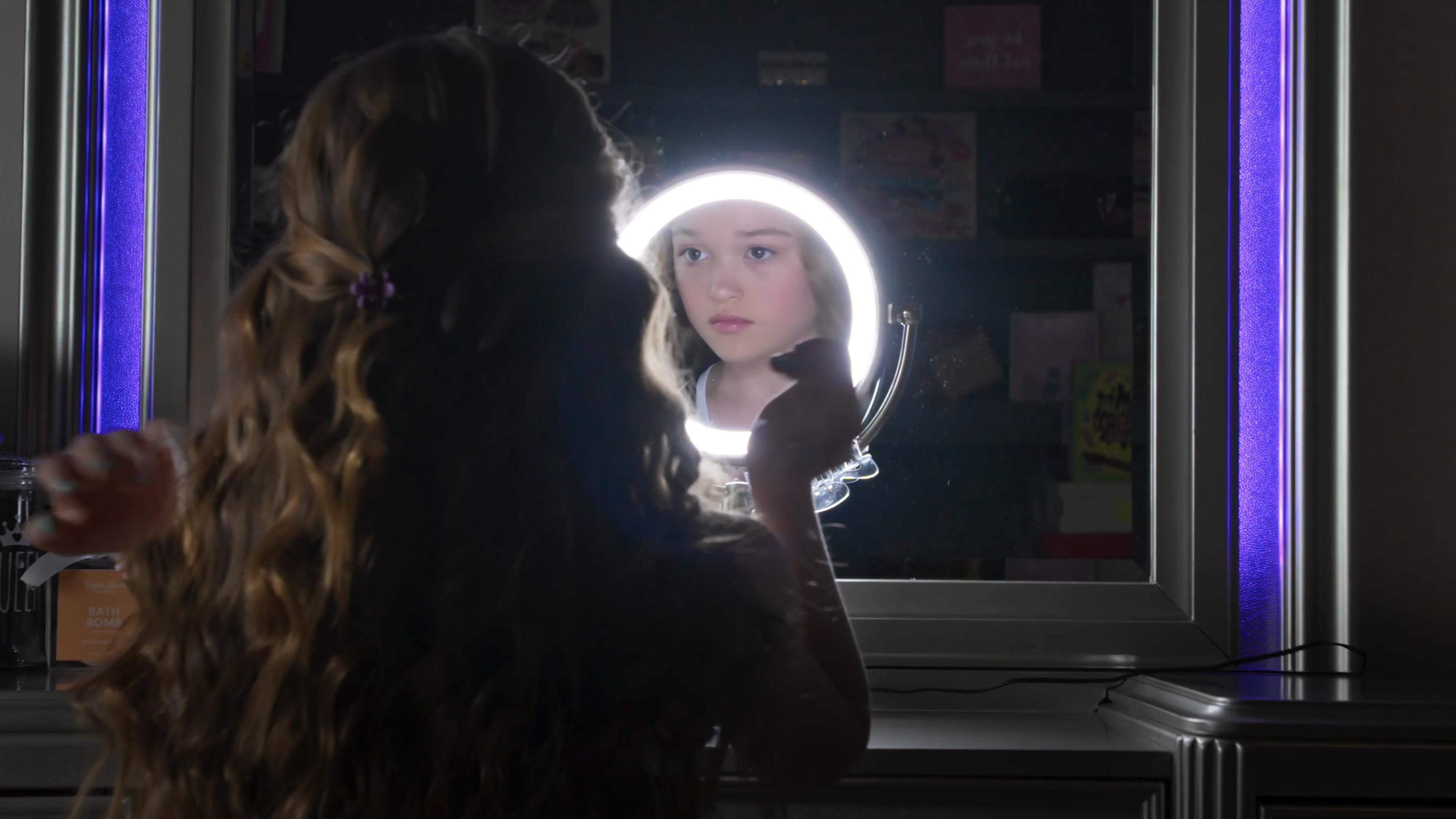 A young girl with curly hair applies makeup in front of a ring-lit mirror, her reflection visible.