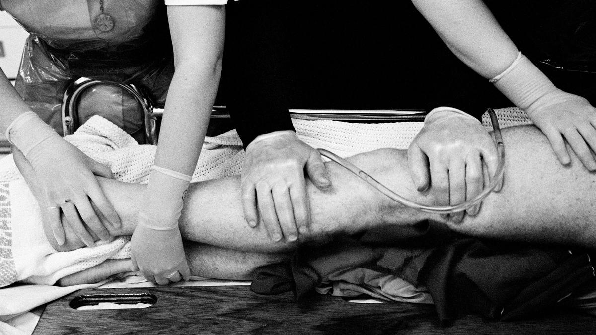 Black and white photo of medical staff handling a patient’s leg wearing gloves, an intravenous line is visible.