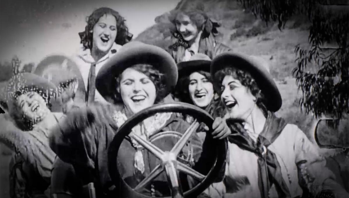 A vintage photo of six women wearing wide-brimmed hats, smiling and laughing together. One woman is holding a steering wheel, suggesting they might be on a fun outdoor adventure. The background shows a natural, slightly blurry setting.