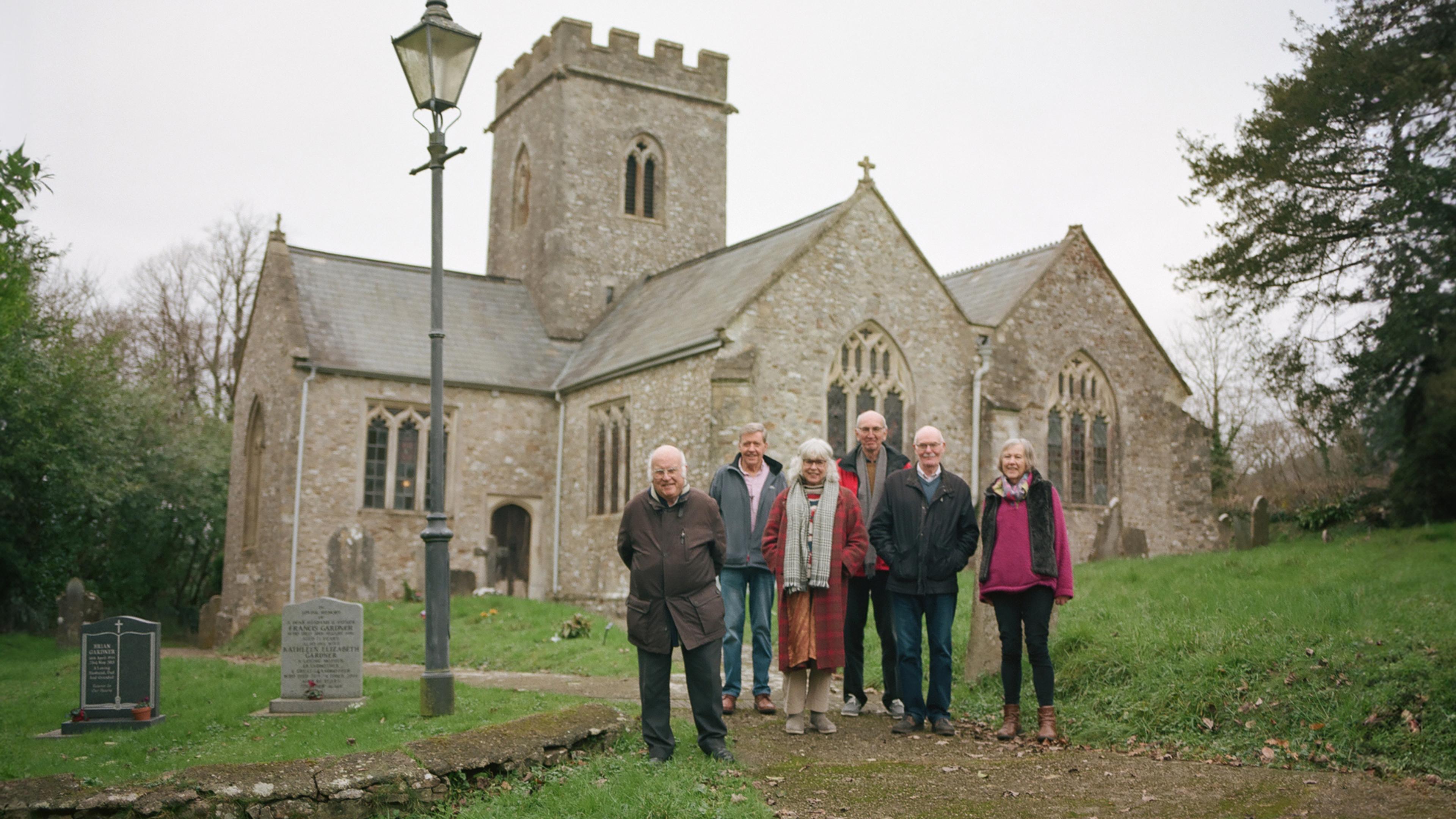 Seven elderly people stand in front of an old church surrounded by a graveyard under a grey sky with trees in the background.