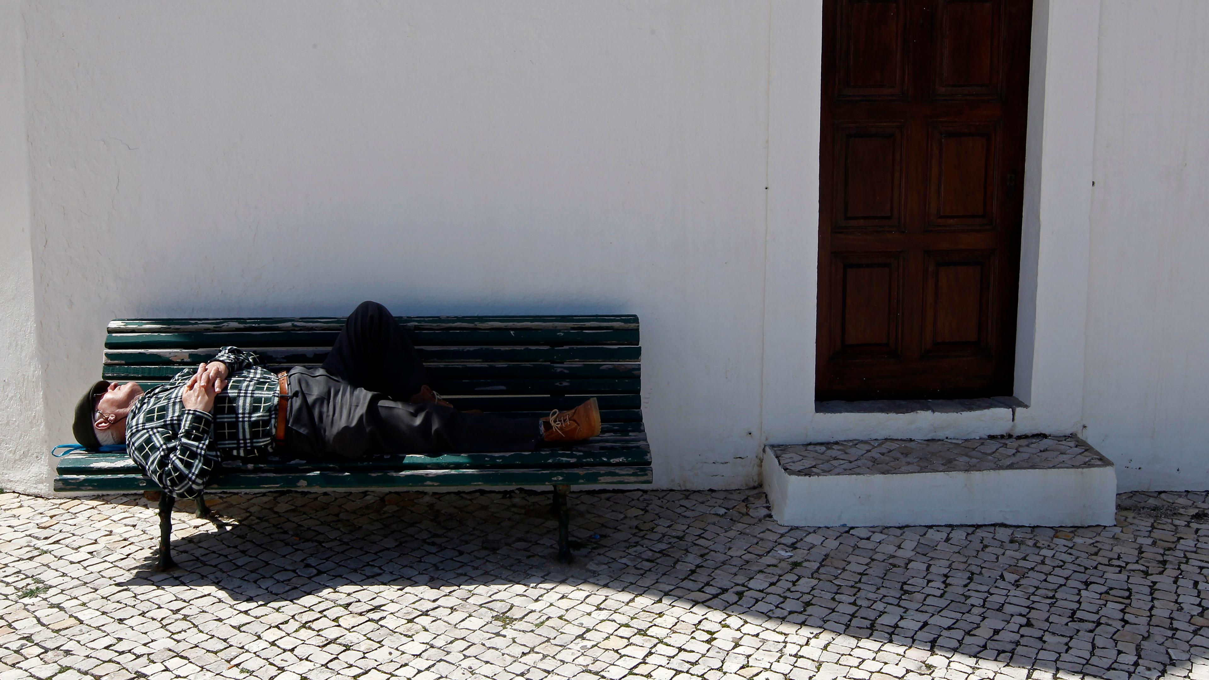 Photo of a man sleeping on a bench against a white wall near a wooden door, with cobblestone paving below in sunlight.