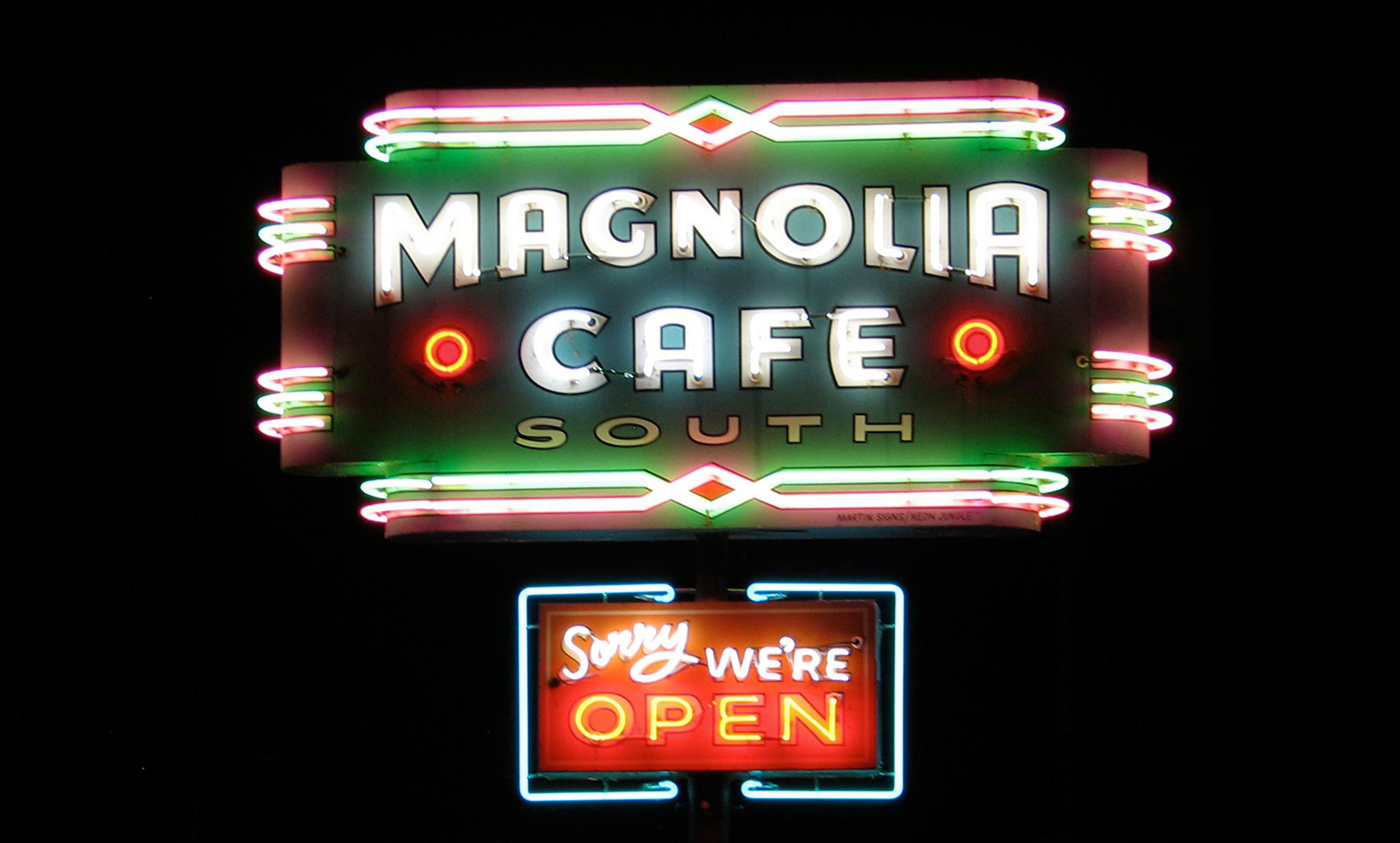 Neon sign for Magnolia Cafe South with colourful lights against a dark background. Below it reads “Sorry we’re open.”
