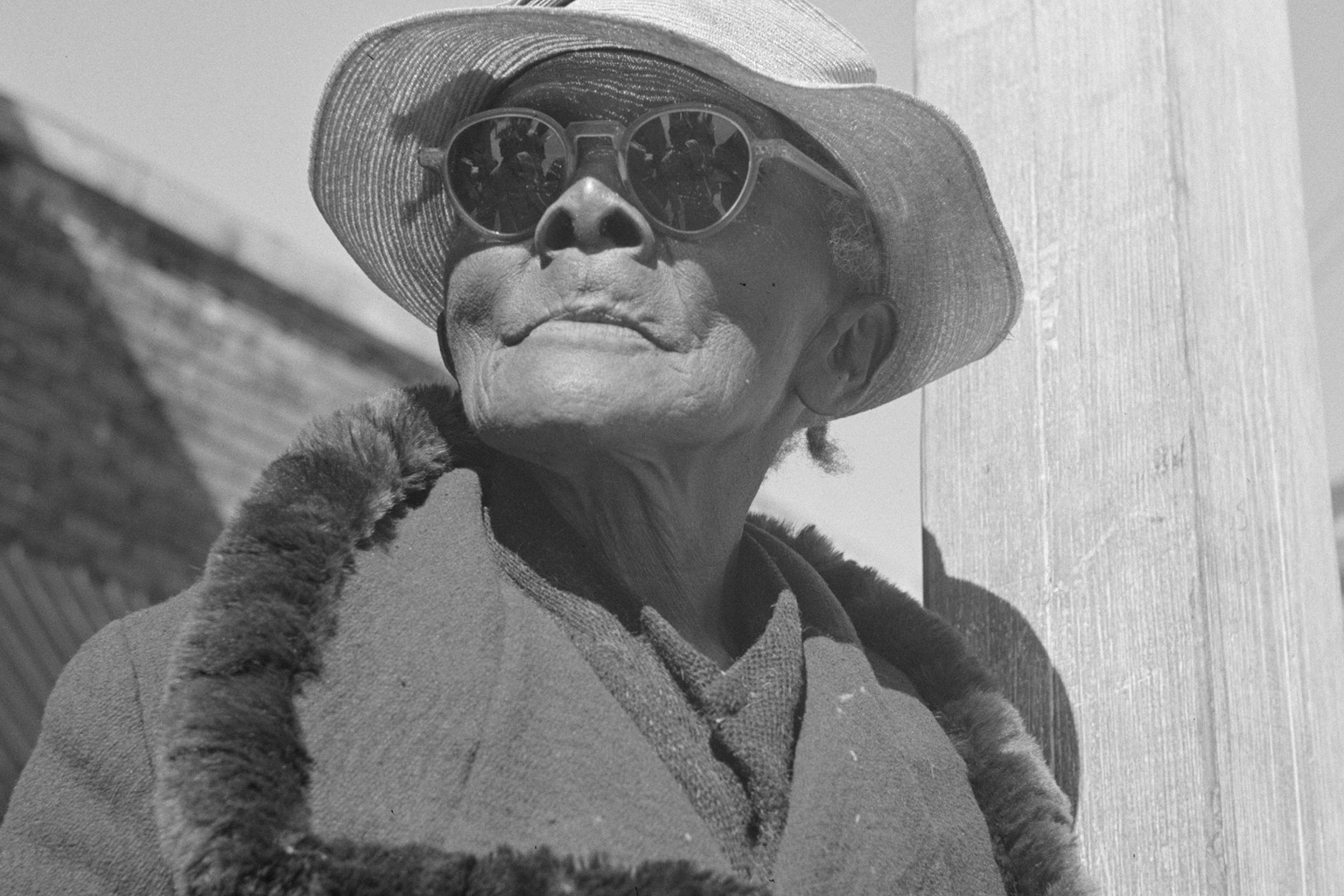 Black and white photo of an elderly person in sunglasses and hat, wearing a fur-collared coat, standing in a street.