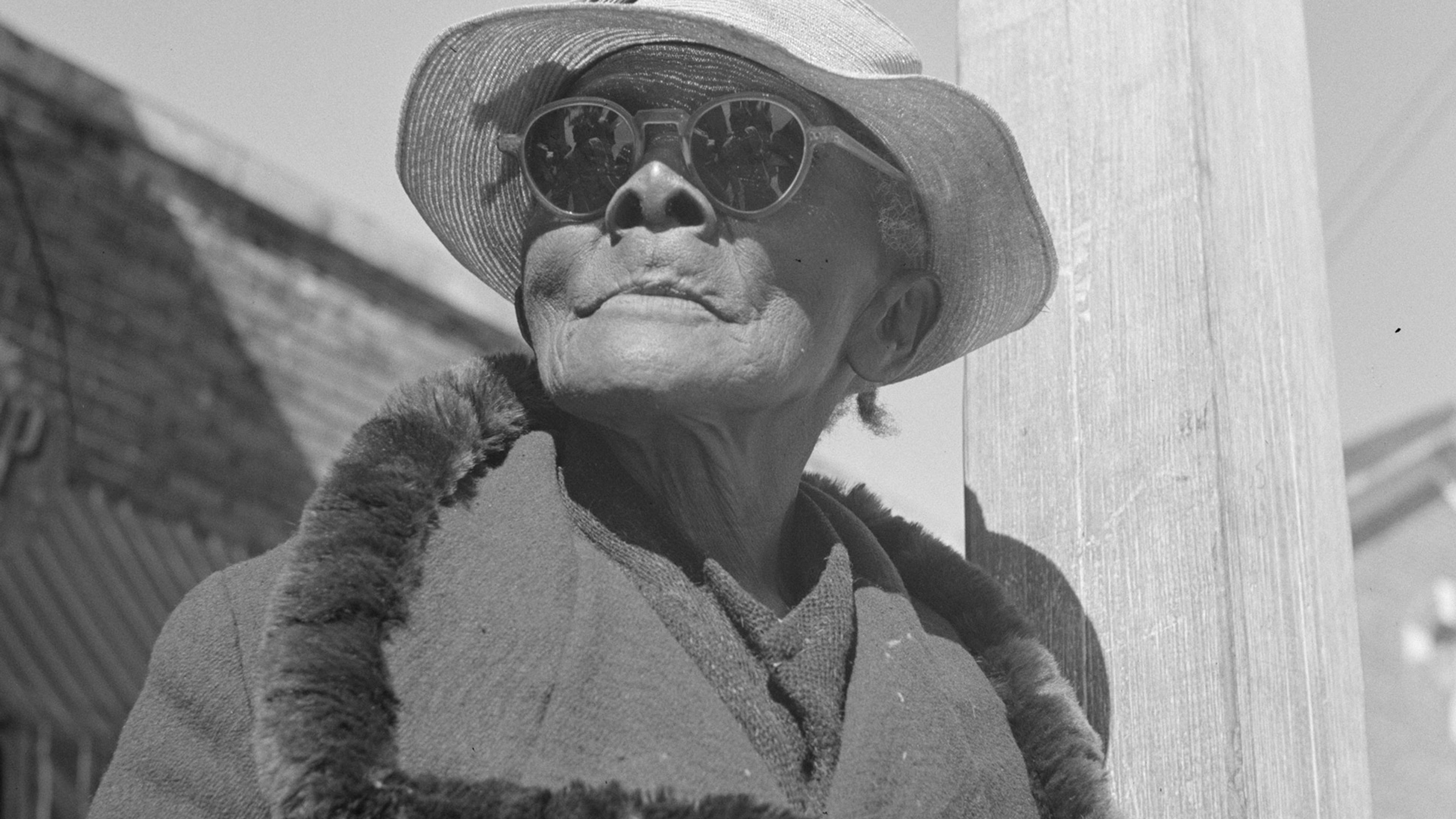 Black and white photo of an elderly person in sunglasses and hat, wearing a fur-collared coat, standing in a street.