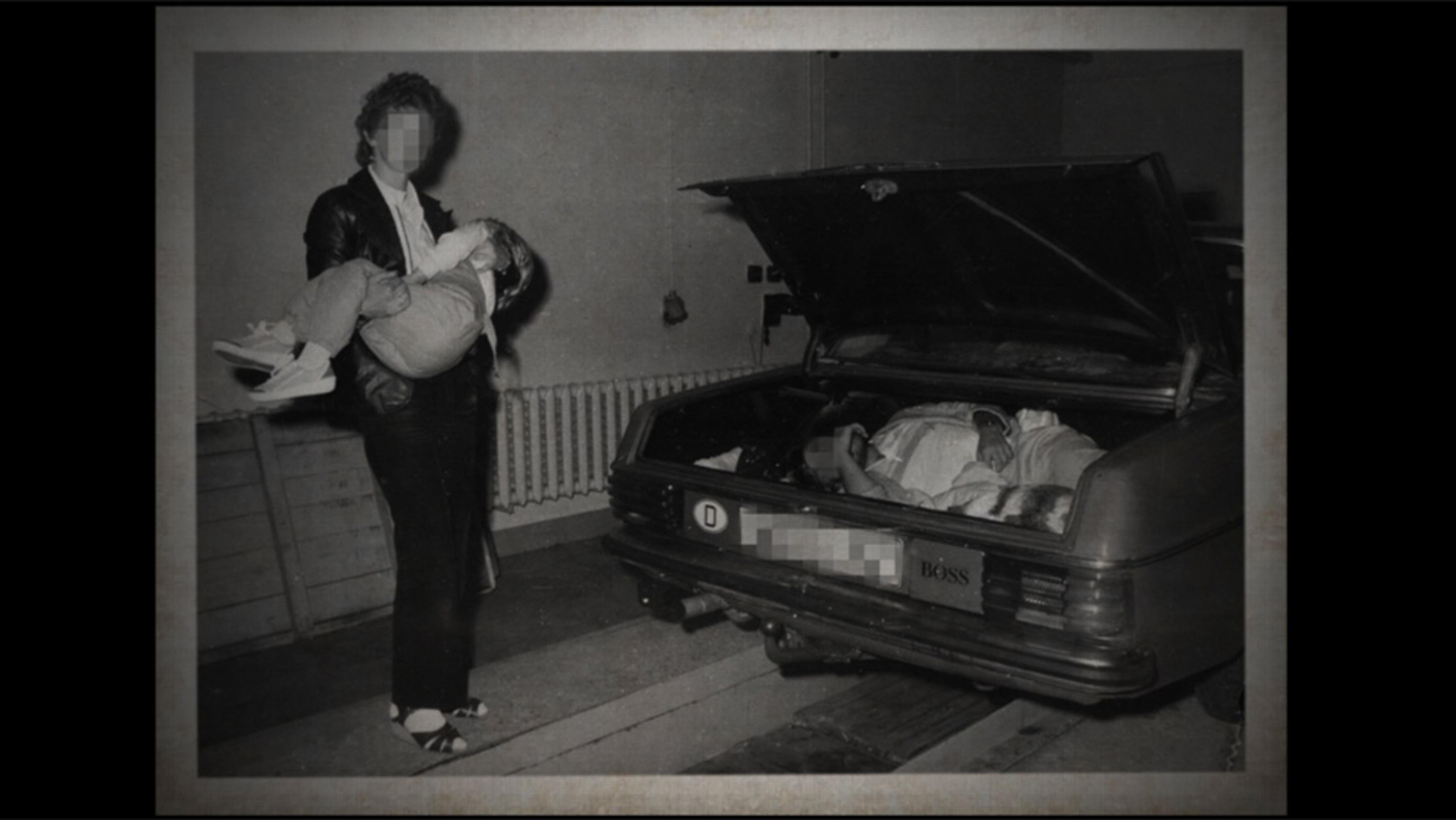 Vintage photo of a woman with a pixelated face holding a sleeping child near a car with an open boot containing blankets or clothes in a dimly lit room.