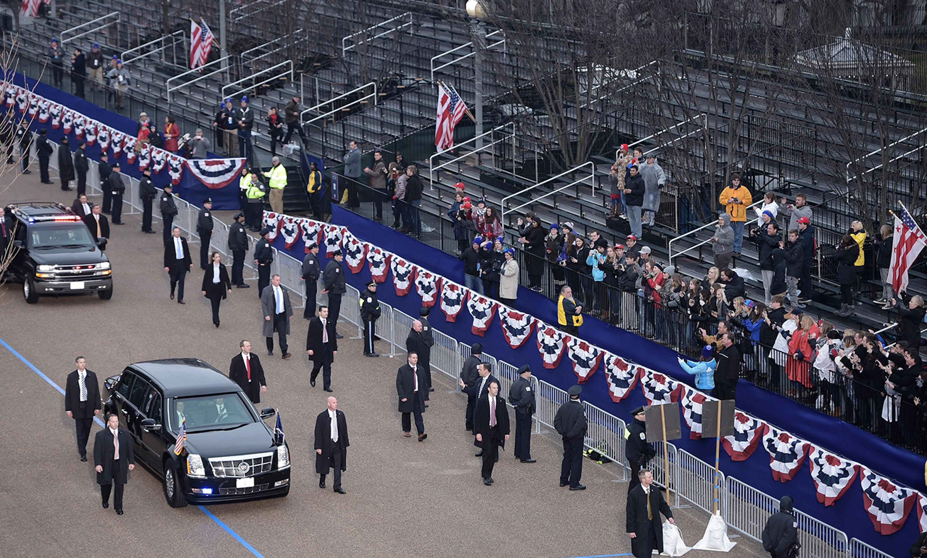 A motorcade driving down a street with bunting; security personnel are walking alongside, and spectators are in stands.
