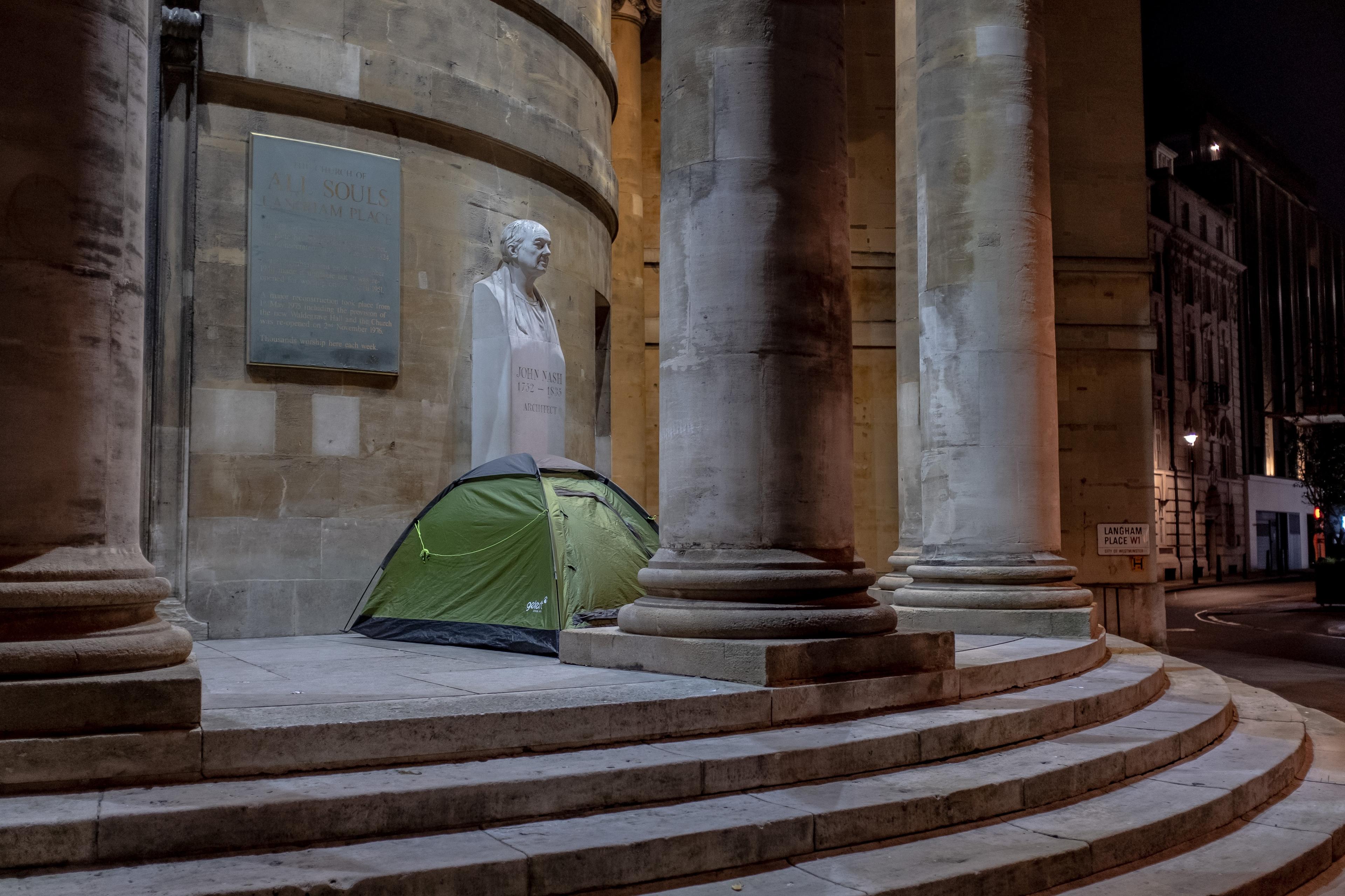 Photo of a green tent pitched on stone steps beside large columns and a bust at night, urban surroundings are visible.