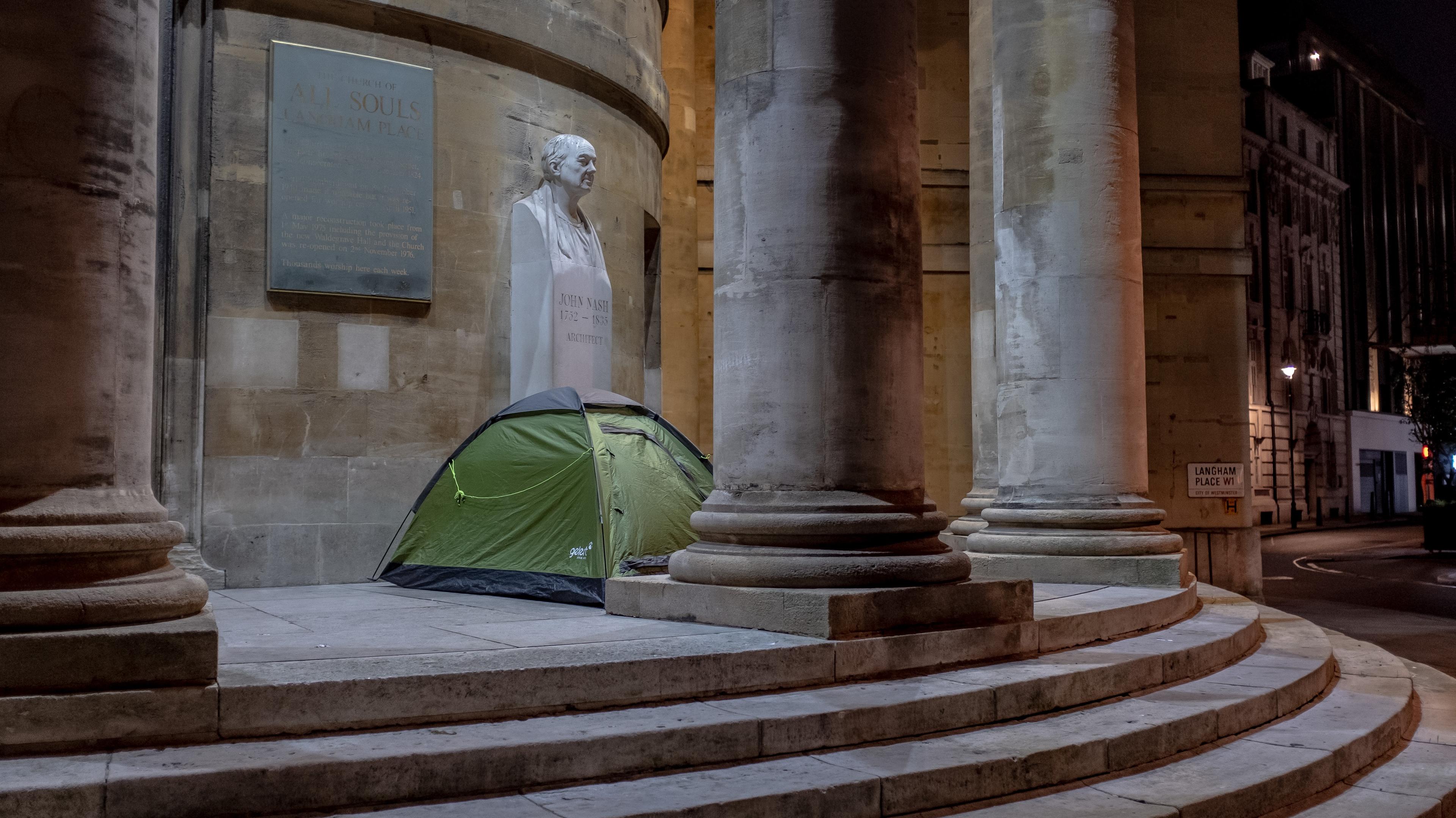 Photo of a green tent pitched on stone steps beside large columns and a bust at night, urban surroundings are visible.