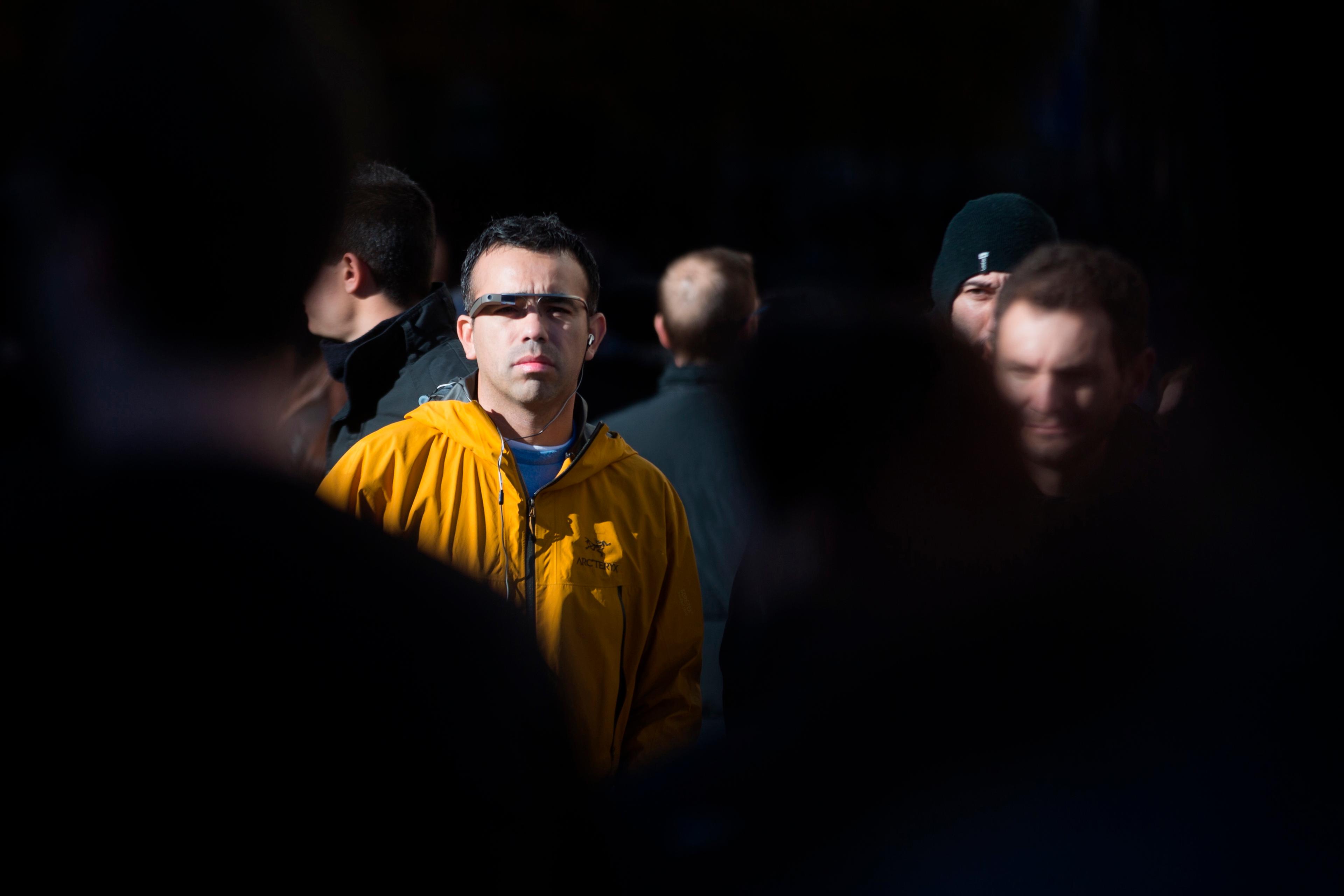 Photo of a man in a yellow jacket wearing smart glasses, standing amidst a dark crowd with light on his face.