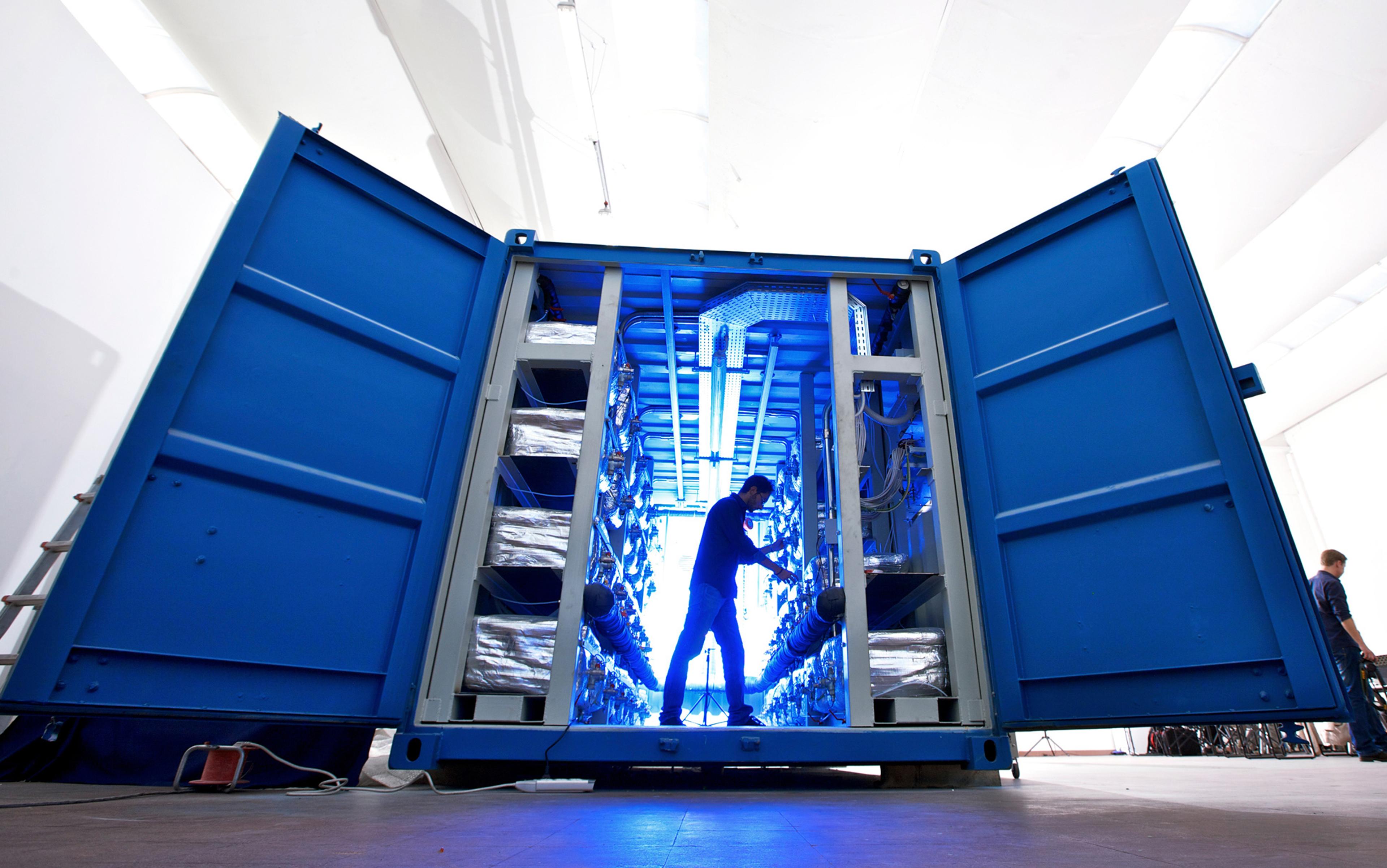 A person working inside a blue industrial container with open doors, showing shelves with equipment, lit by blue light.