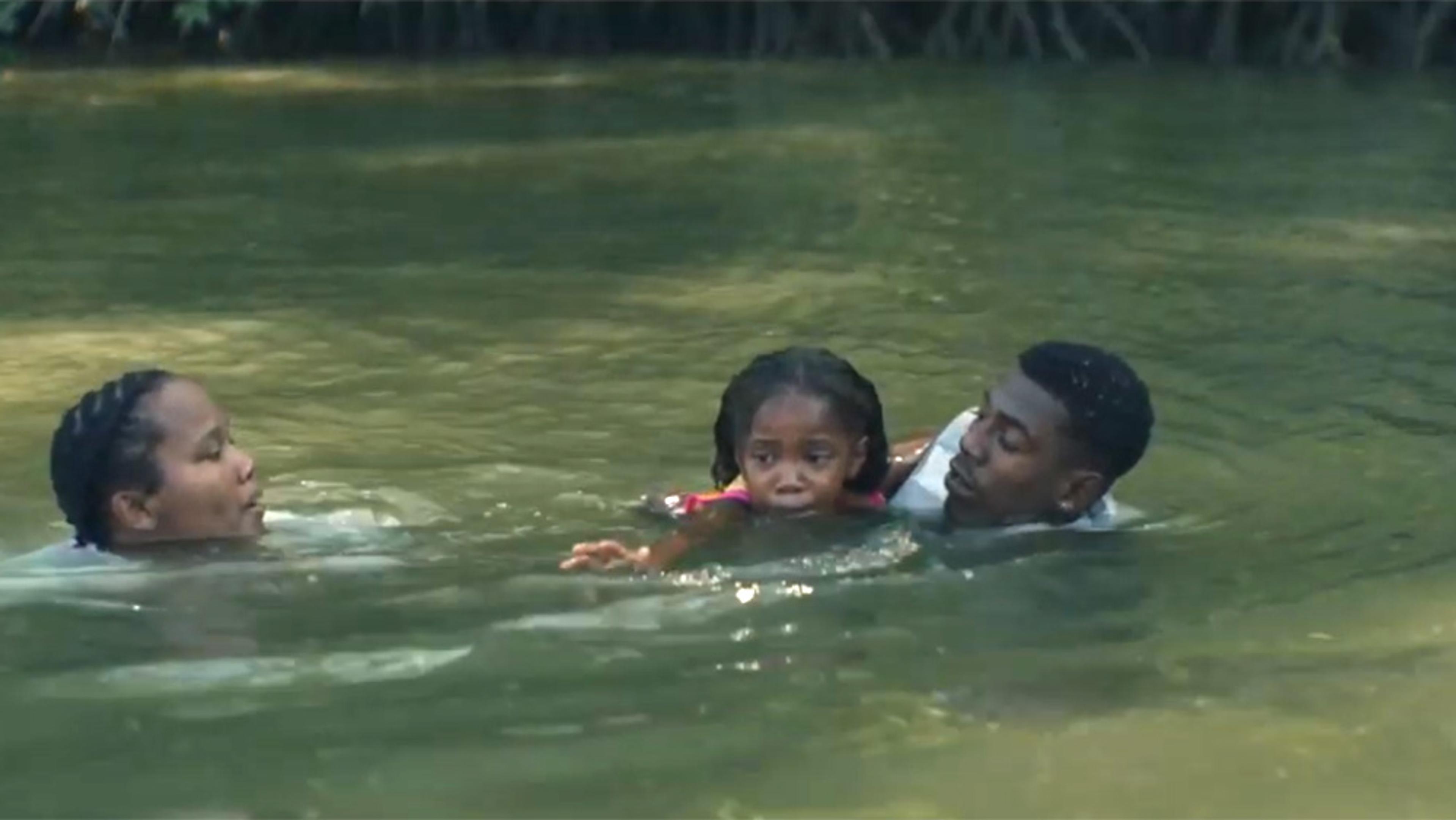 Photo of two adults swimming with a young child in a natural body of water, lush greenery in the background.