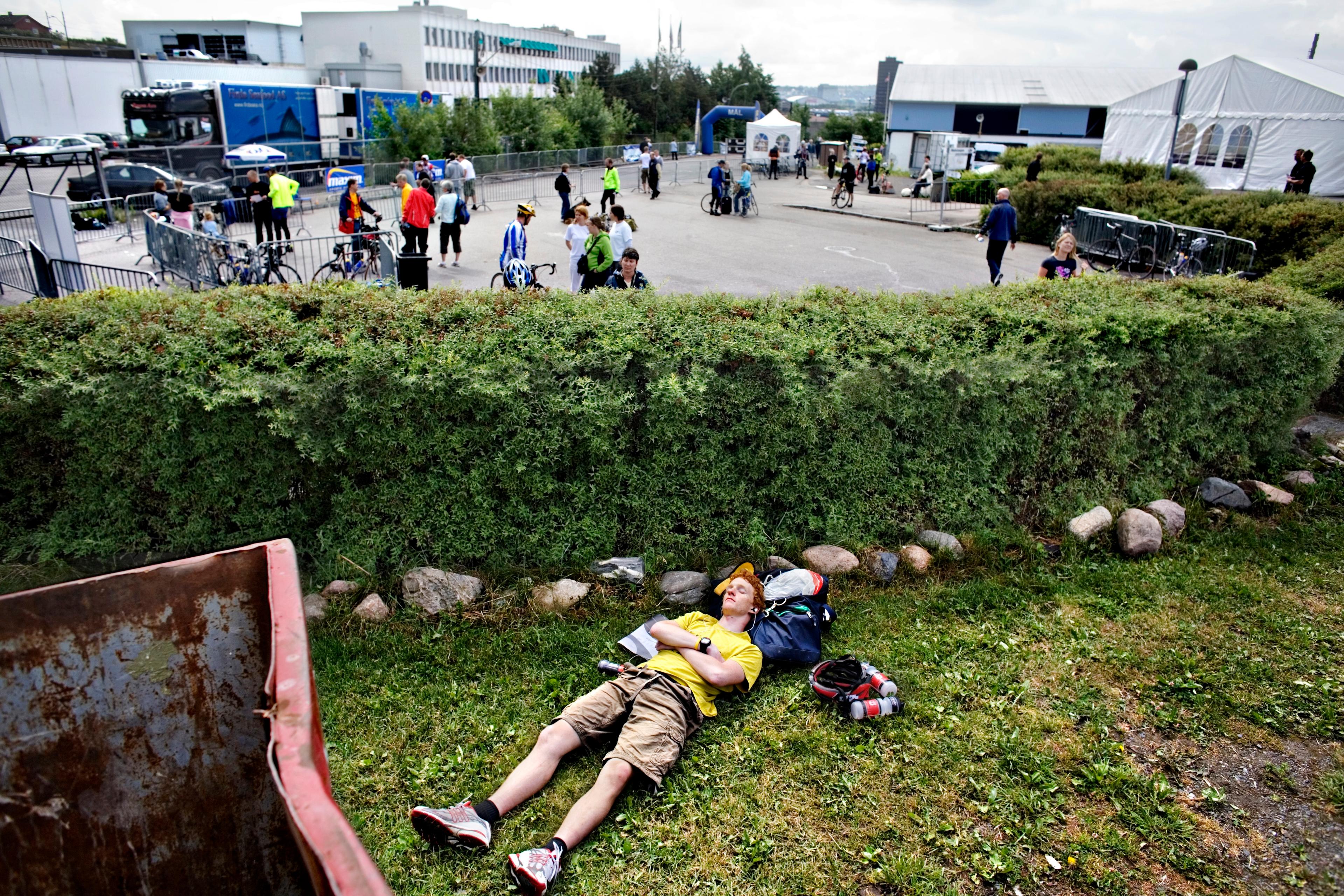 Photo of a man in a yellow shirt resting on grass by a hedge with cyclists and tents in the background.