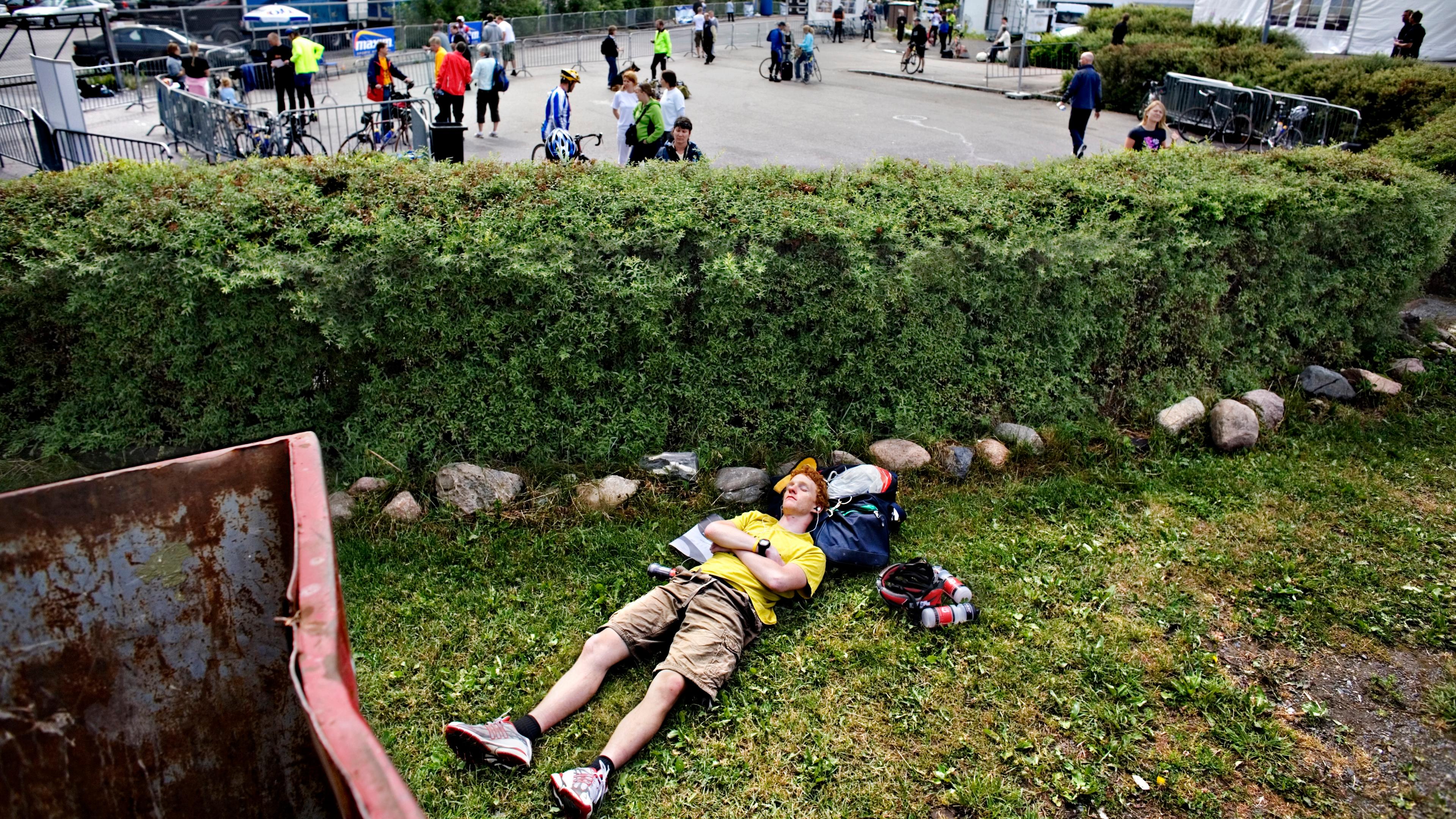 Photo of a man in a yellow shirt resting on grass by a hedge with cyclists and tents in the background.