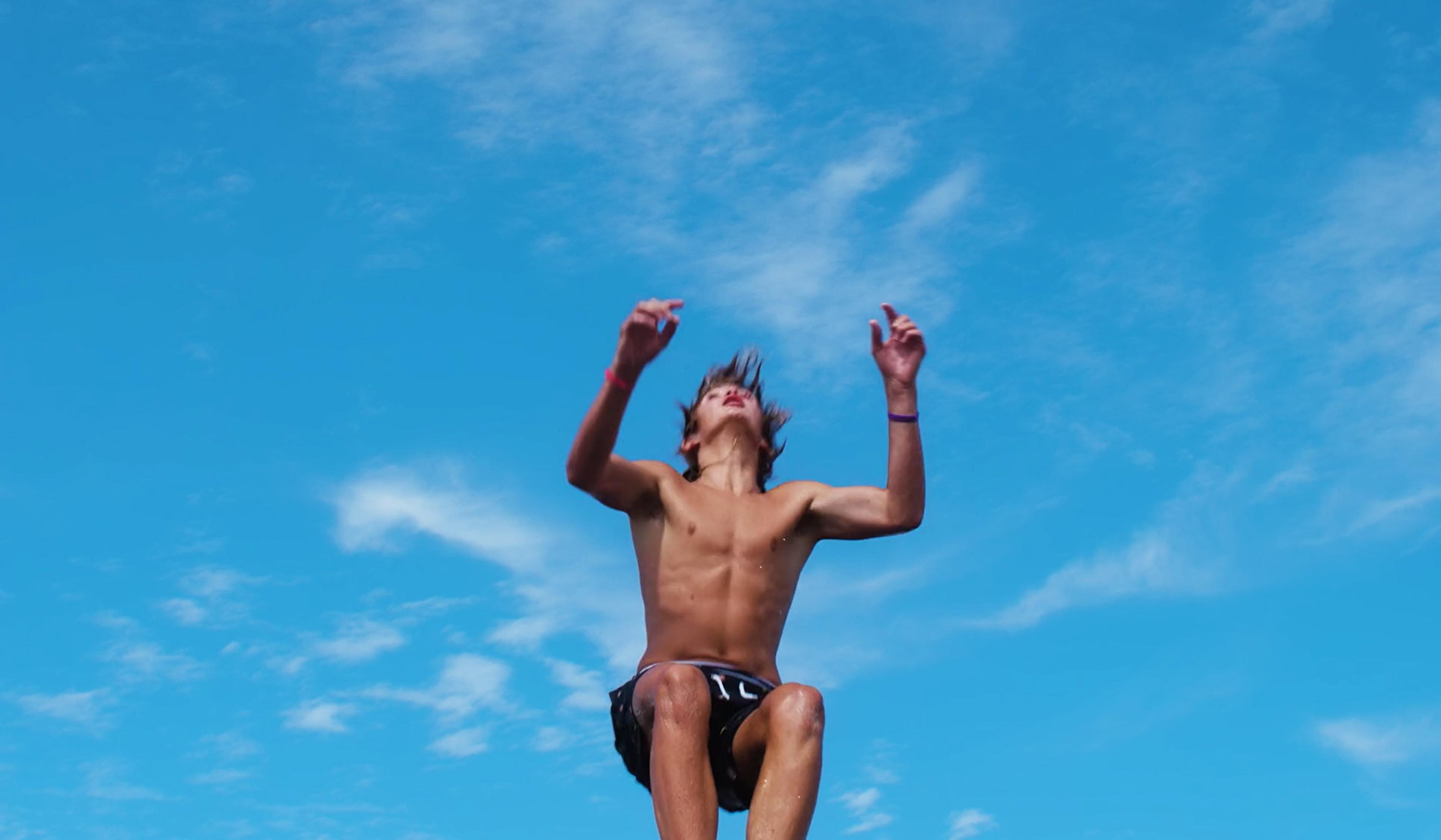 Shirtless youth jumping with arms raised against a blue sky with scattered clouds.