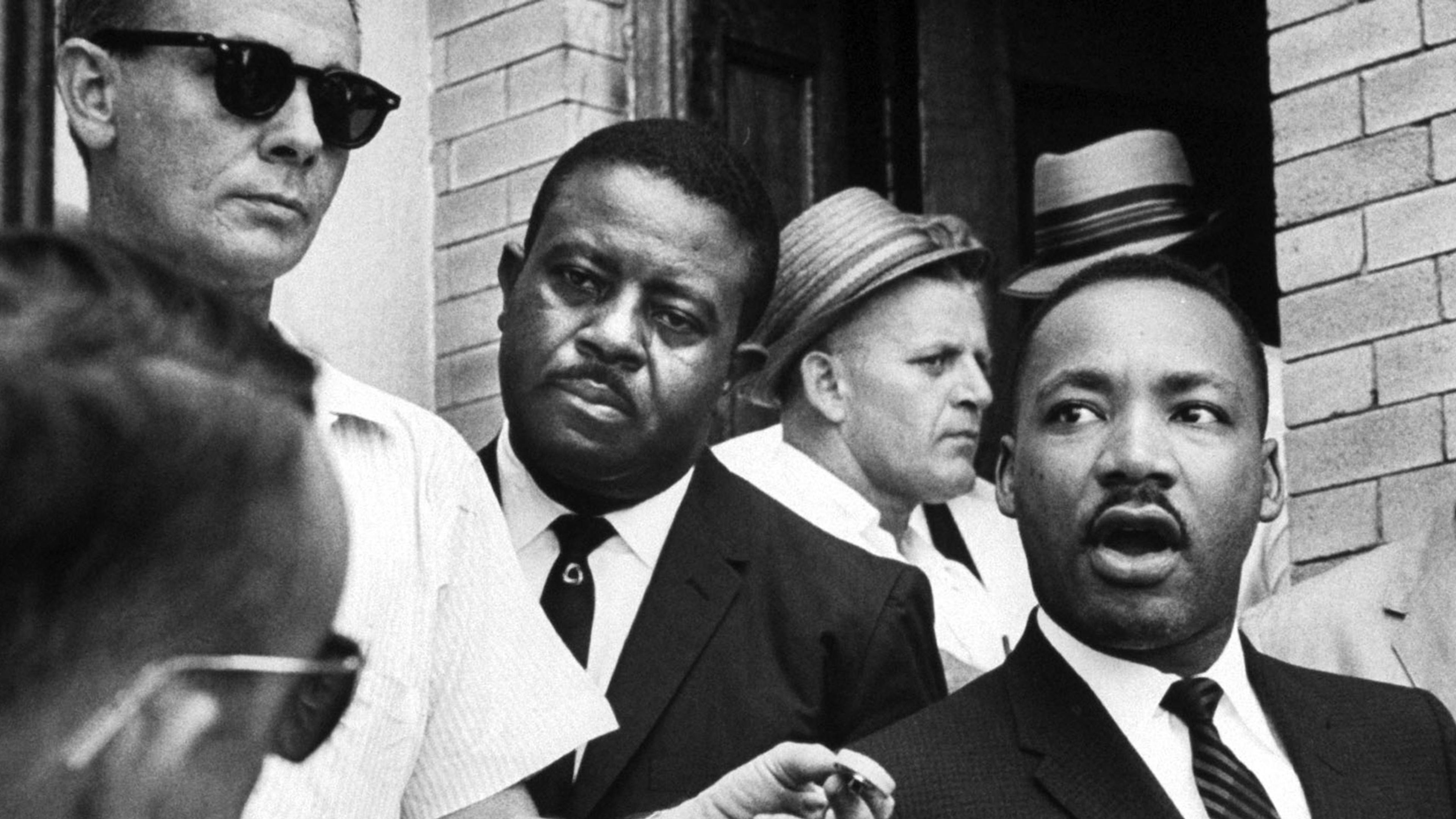 Black-and-white photo of a man speaking to reporters holding microphones in a crowded outdoor setting.
