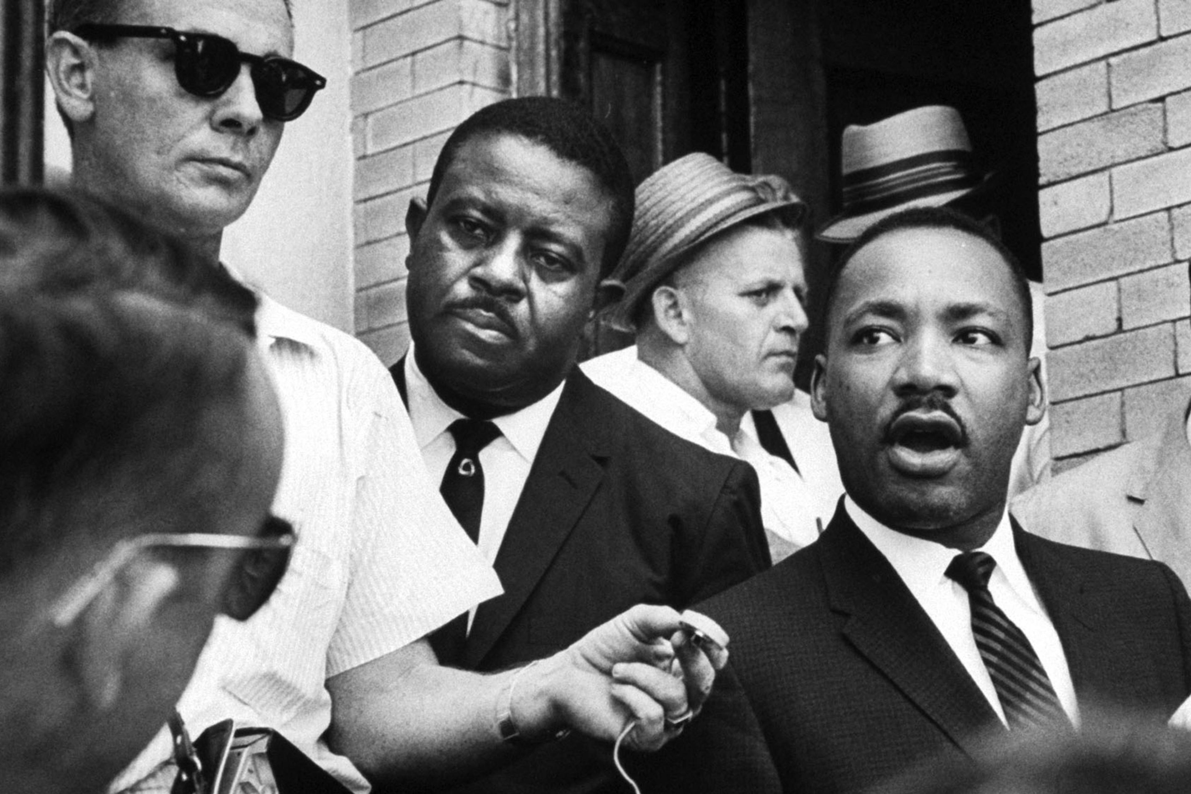 Black-and-white photo of a man speaking to reporters holding microphones in a crowded outdoor setting.
