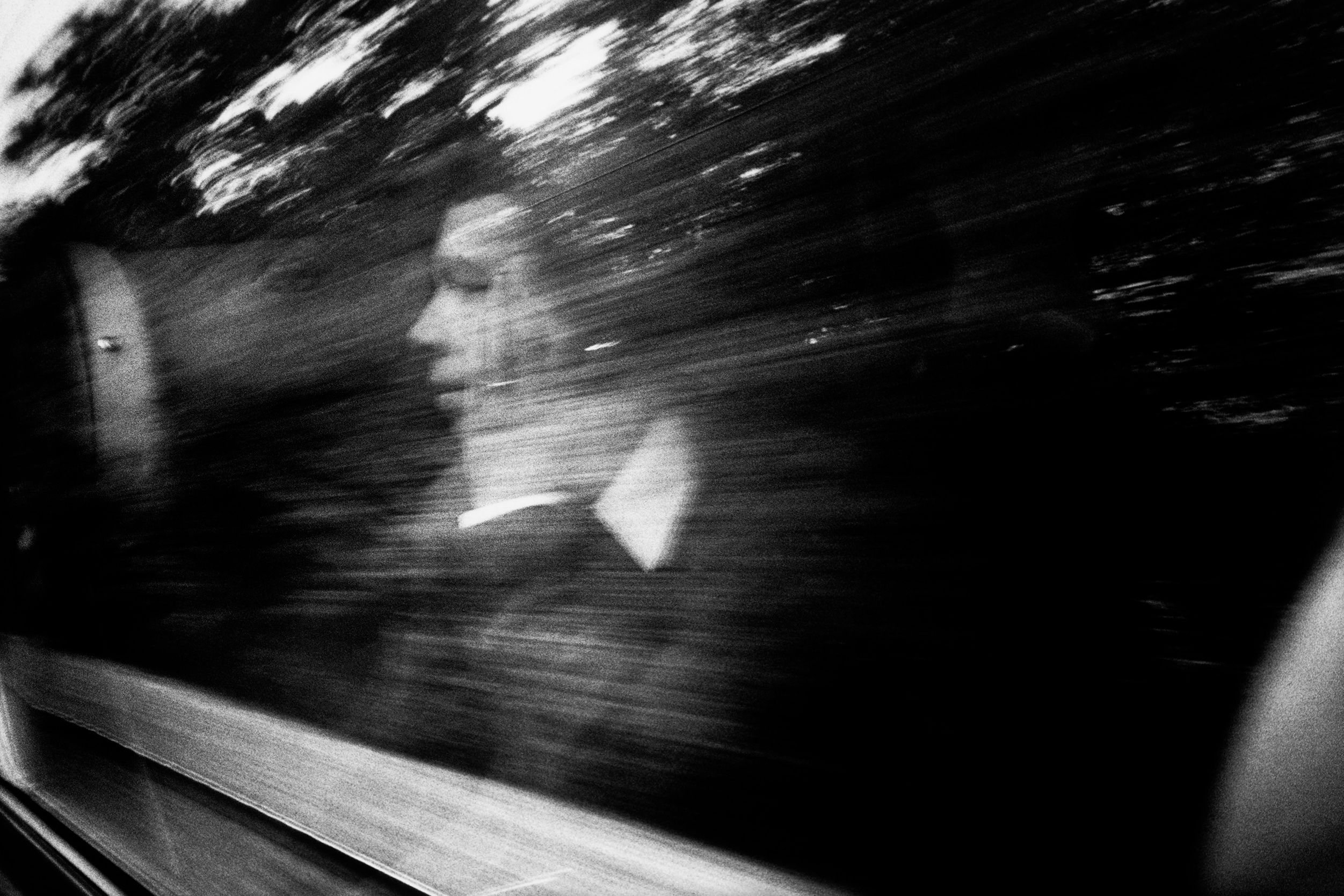 Black and white photo of a person reflected in a window with motion blur and trees visible outside.