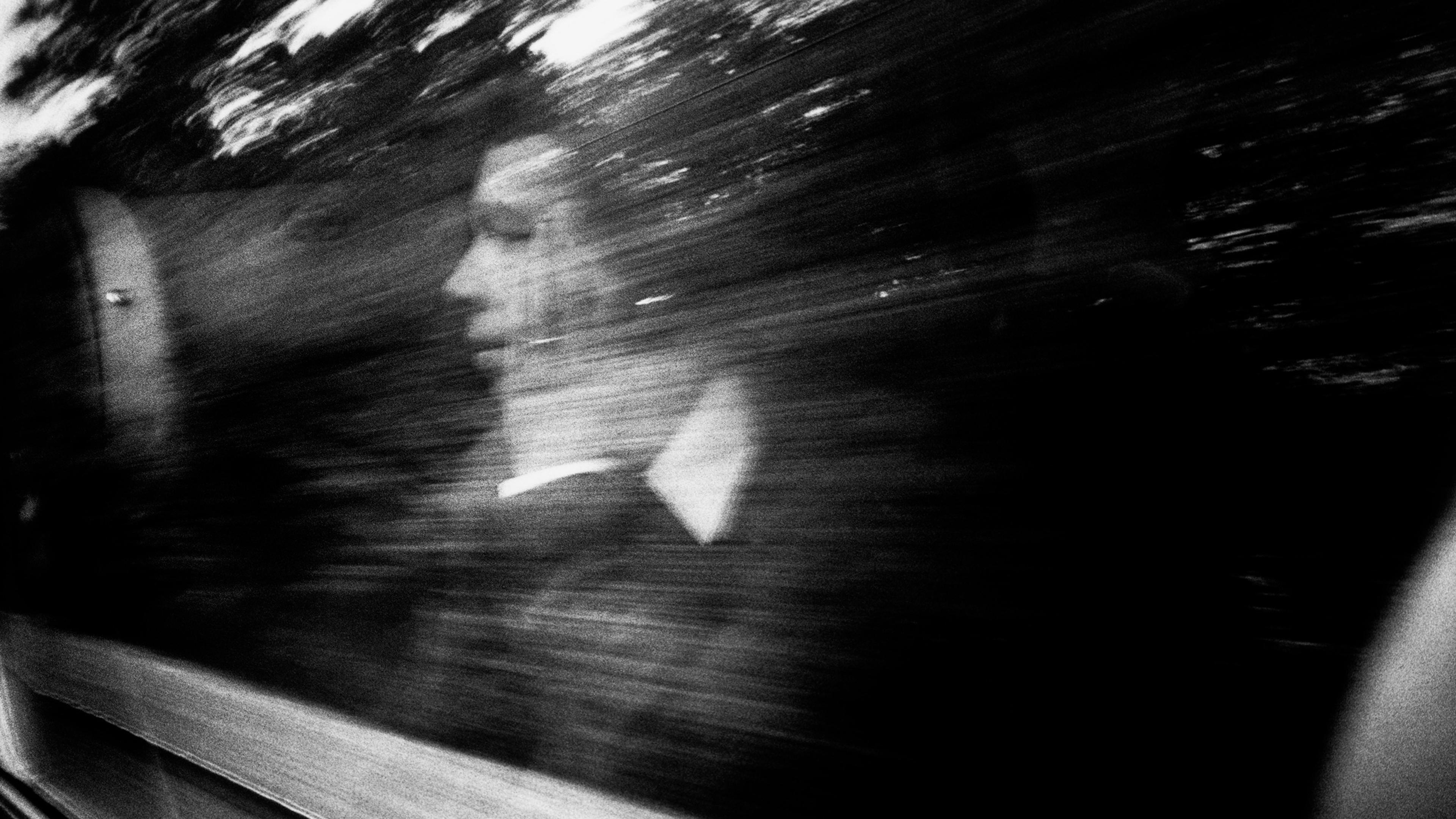 Black and white photo of a person reflected in a window with motion blur and trees visible outside.