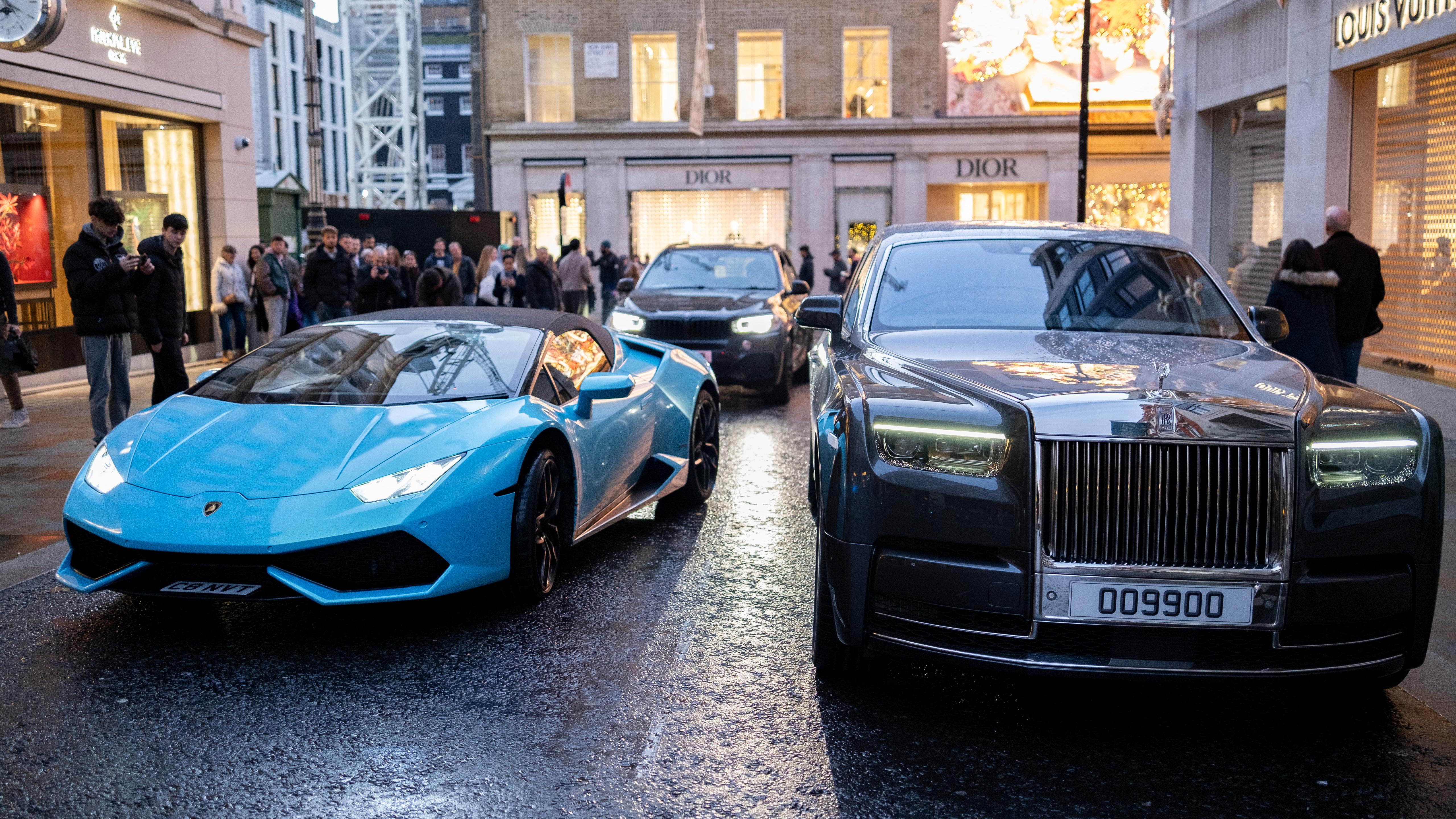 A blue sports car and a grey luxury car parked on a city street with people and a Christian Dior shop in the background.