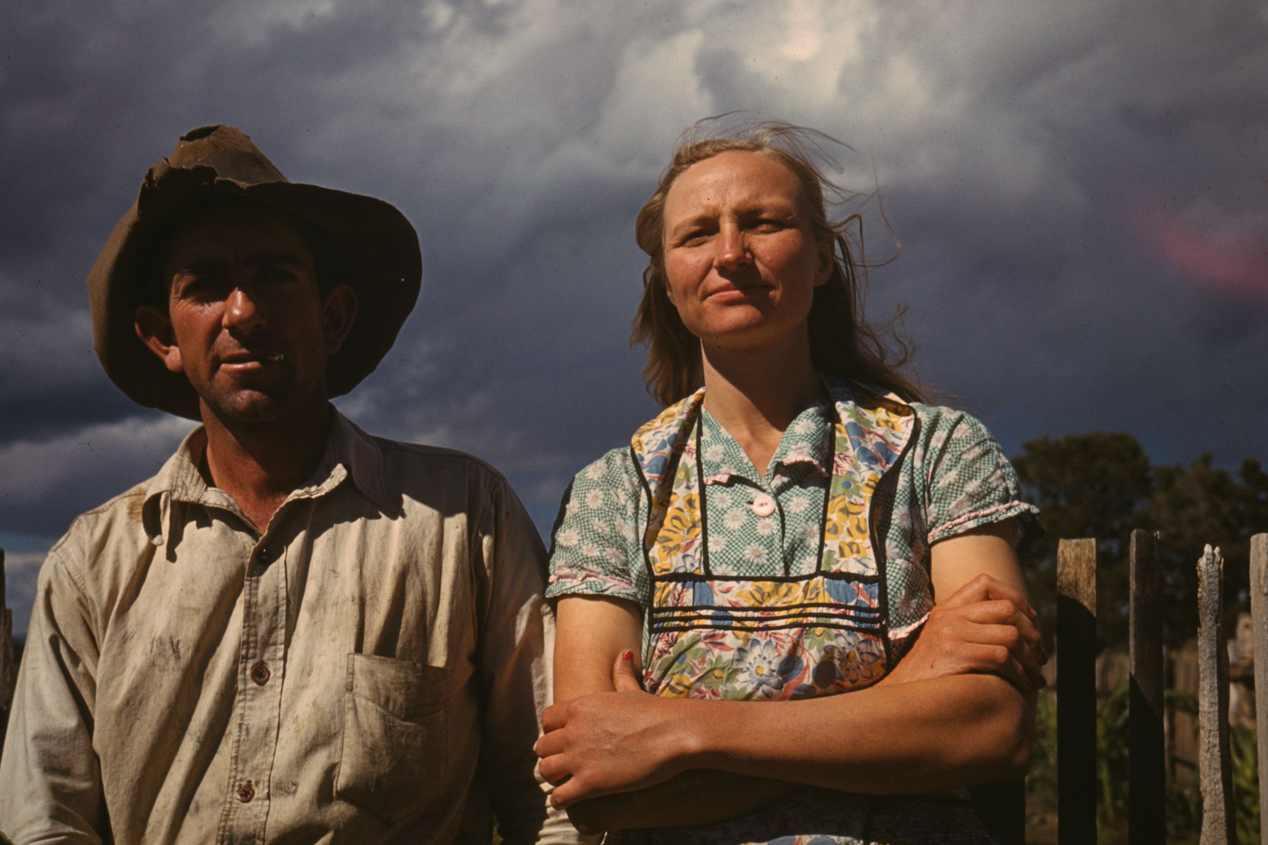Vintage photo of a man in a hat and a woman in a dress standing outside, with a cloudy sky in the background.