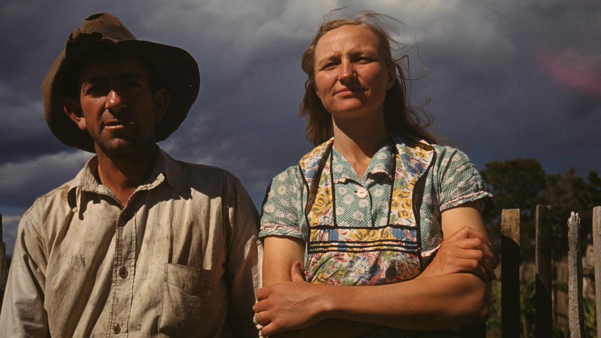 Vintage photo of a man in a hat and a woman in a dress standing outside, with a cloudy sky in the background.