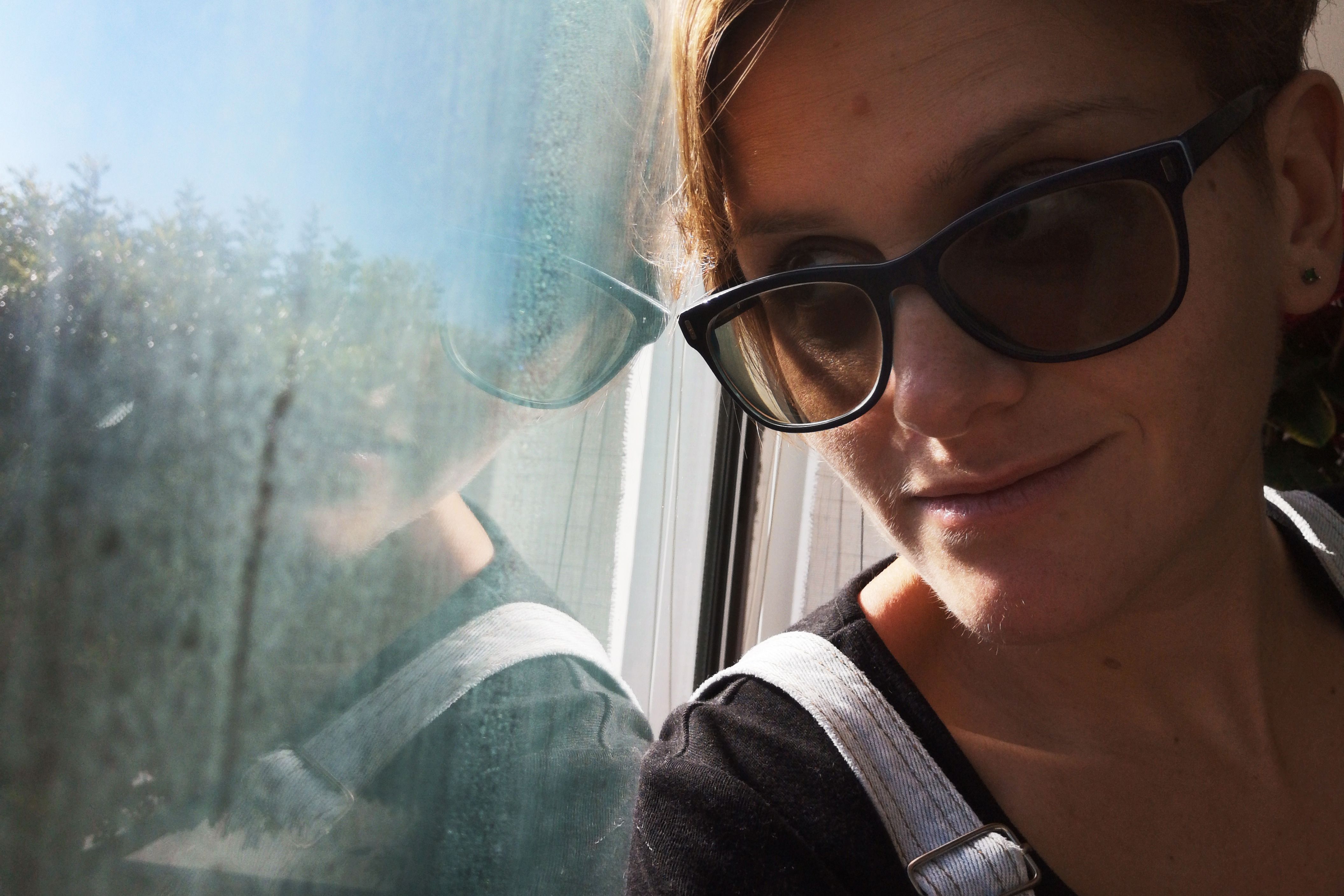A person wearing sunglasses looking at their reflection in a window with a blurry outdoor scene visible.