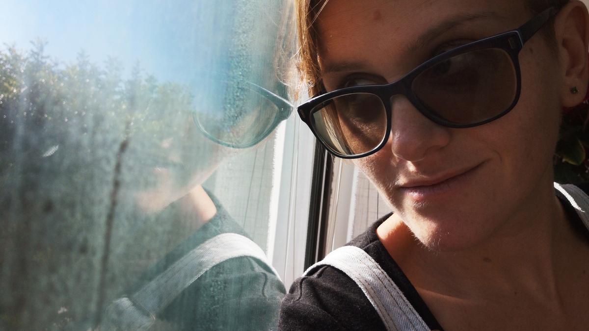 A person wearing sunglasses looking at their reflection in a window with a blurry outdoor scene visible.