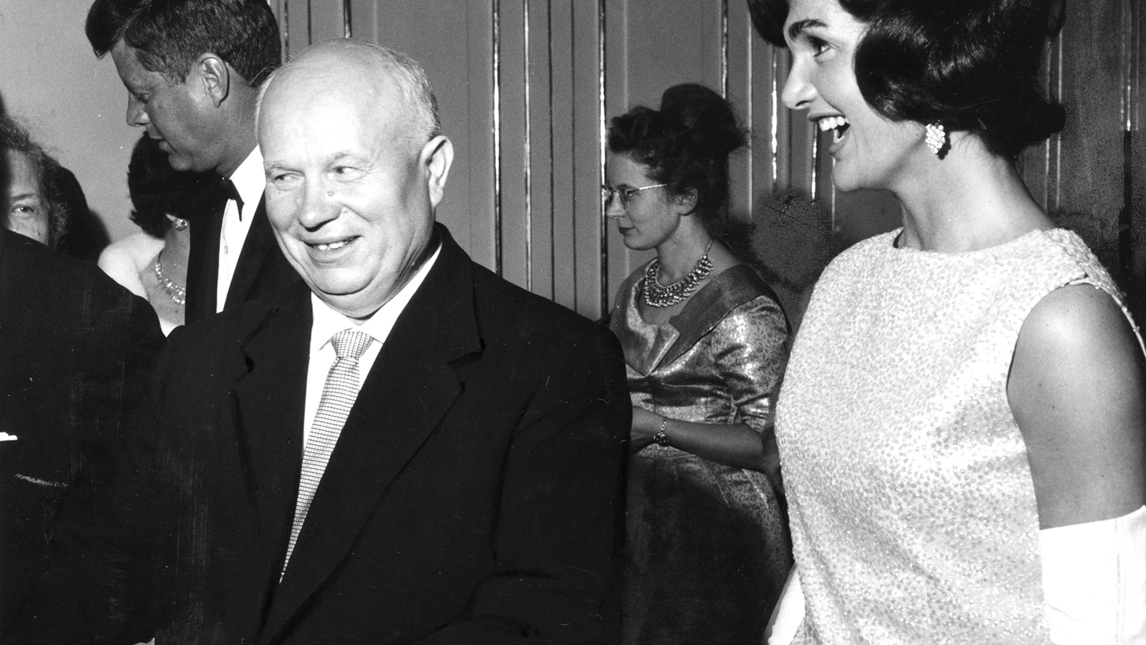 Black and white photo of a man smiling next to a woman laughing at a formal event. Other guests visible in the background.