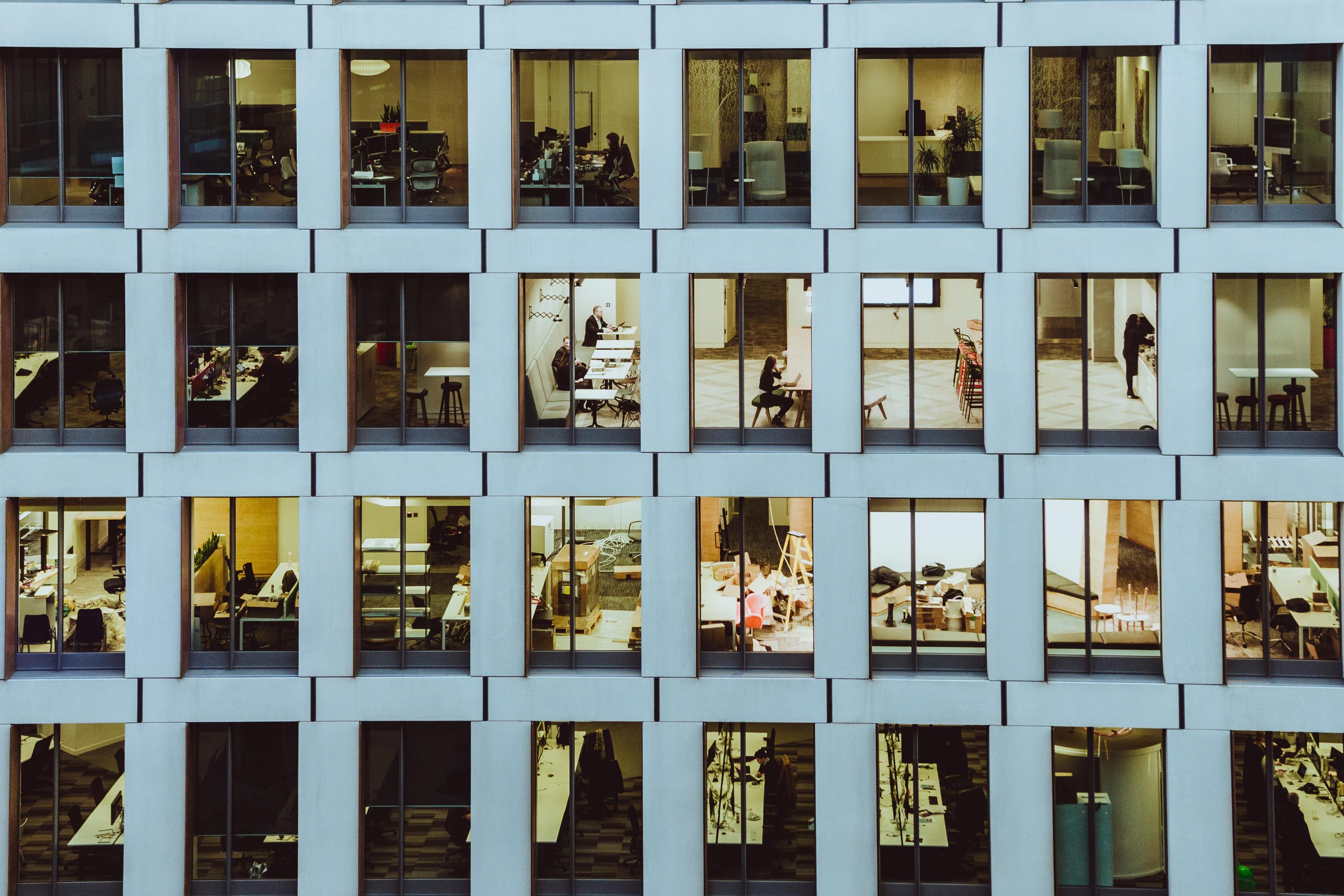 Photo of an office building’s facade with illuminated windows revealing people working inside at desks and meeting spaces.