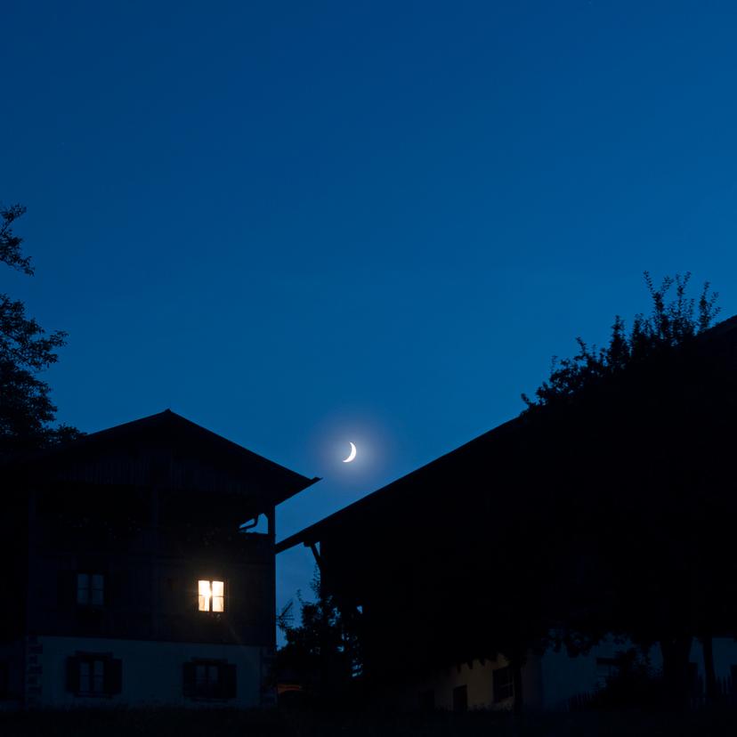 Houses at night with a crescent moon in a clear blue sky, one house has an illuminated window, tree silhouettes frame the scene.