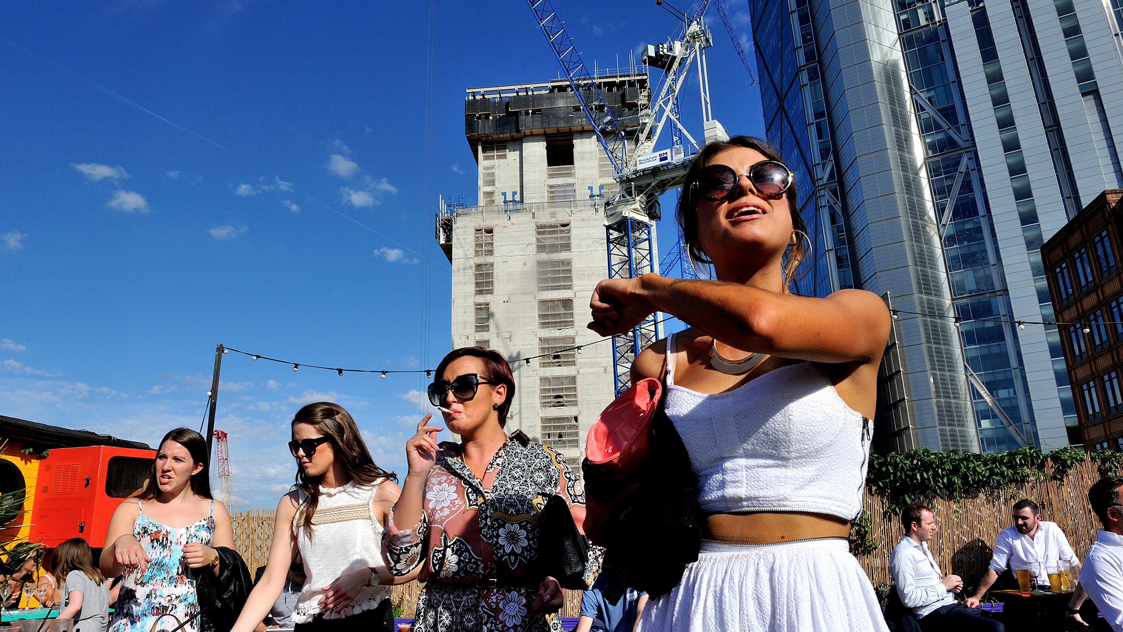 Four women outside wearing summer outfits with skyscrapers and a construction crane in the background; one woman is shown smoking.