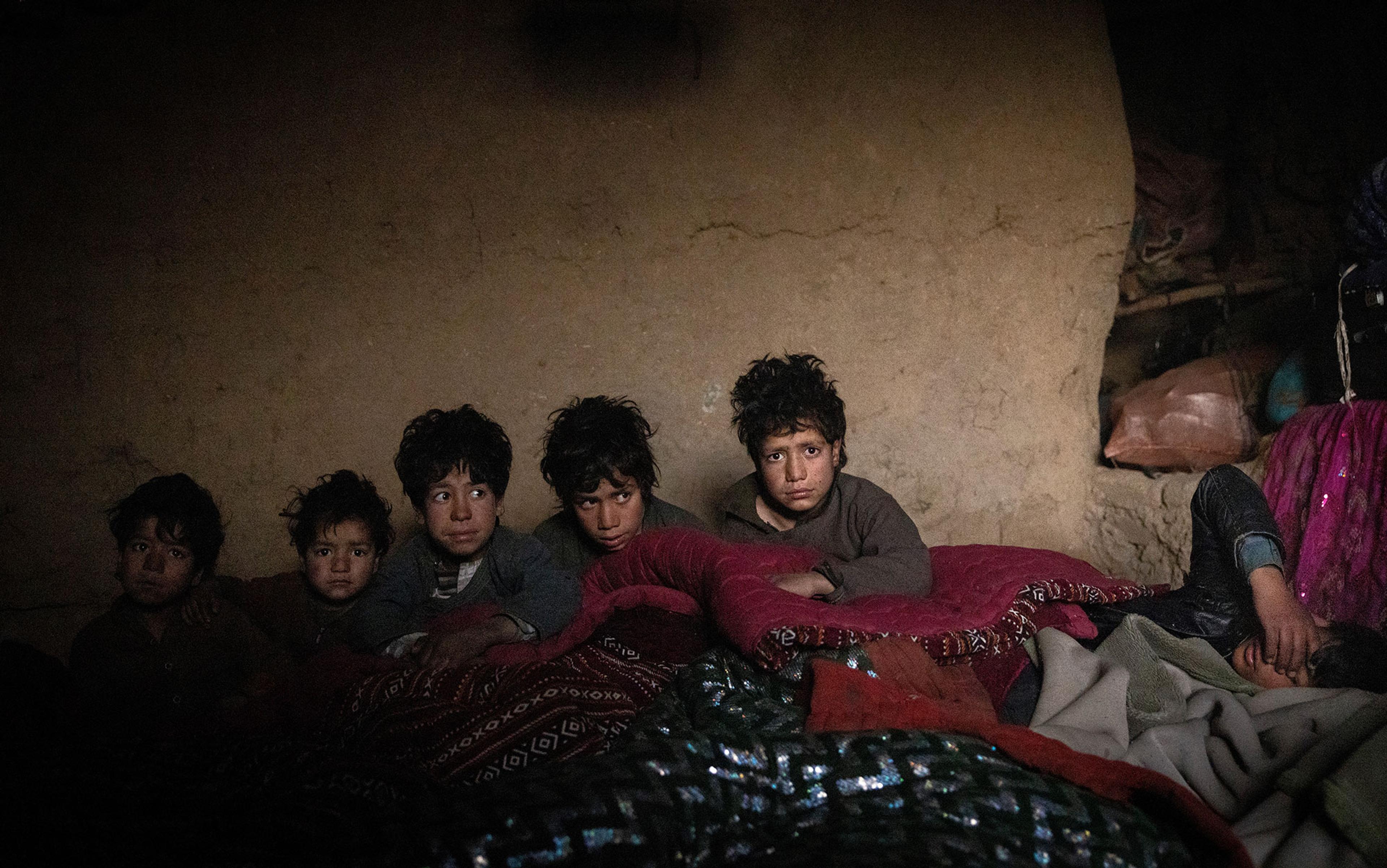 Six children with troubled faces sitting and lying on colourful blankets inside a dimly lit room with an earthen wall.