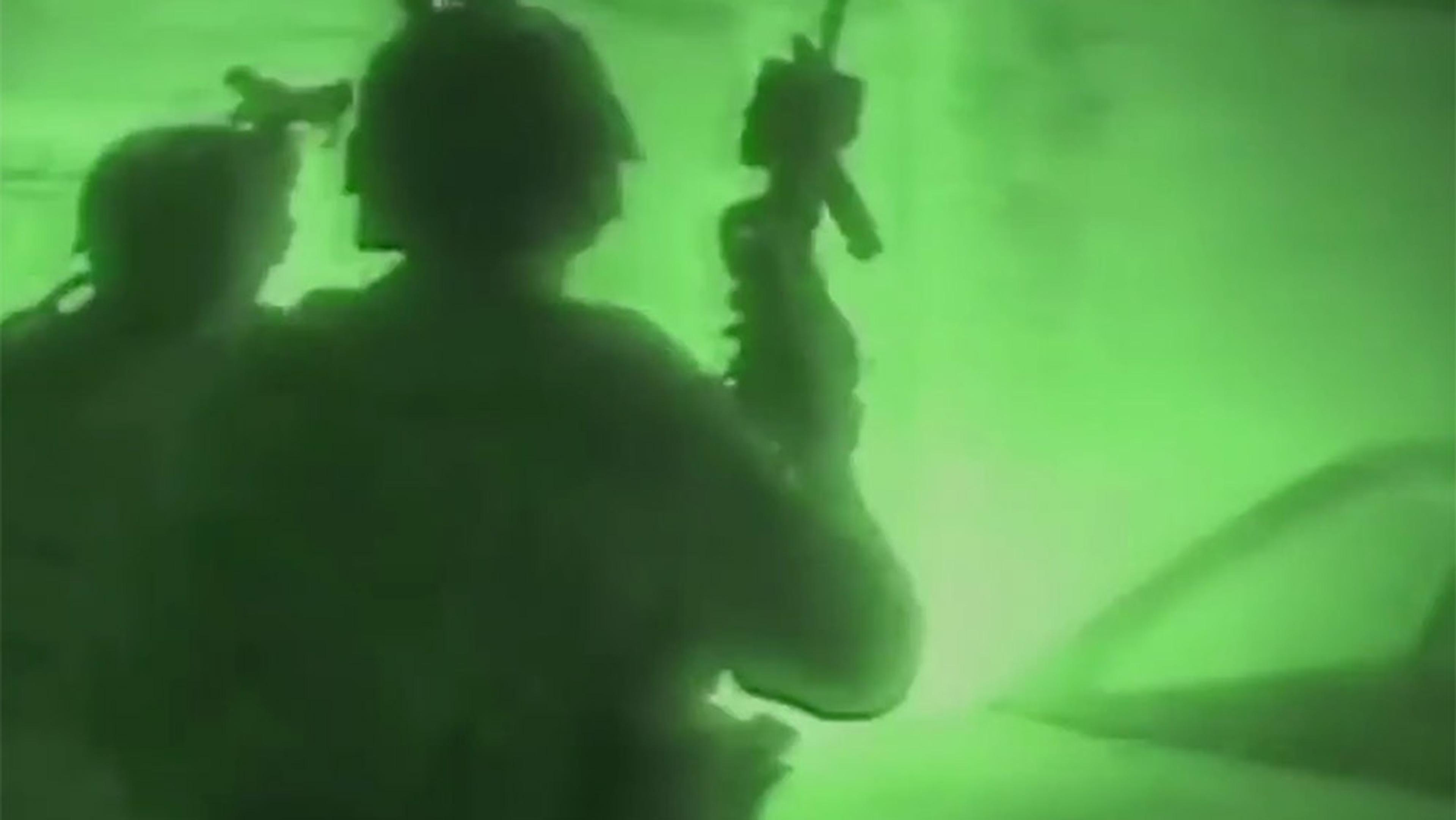Ttwo soldiers seen through green night vision goggles, holding rifles in a dimly lit environment with a vehicle in view.