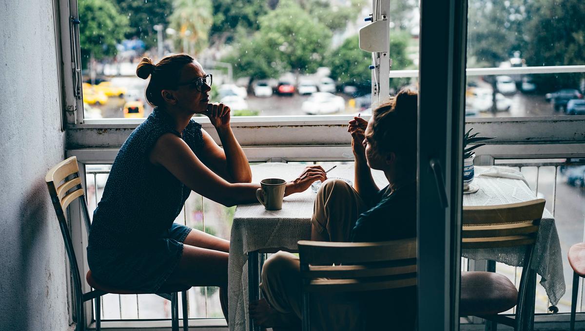 Two people sit at a table with coffee cups, near a window overlooking a rainy scene with parked cars and trees in the background.