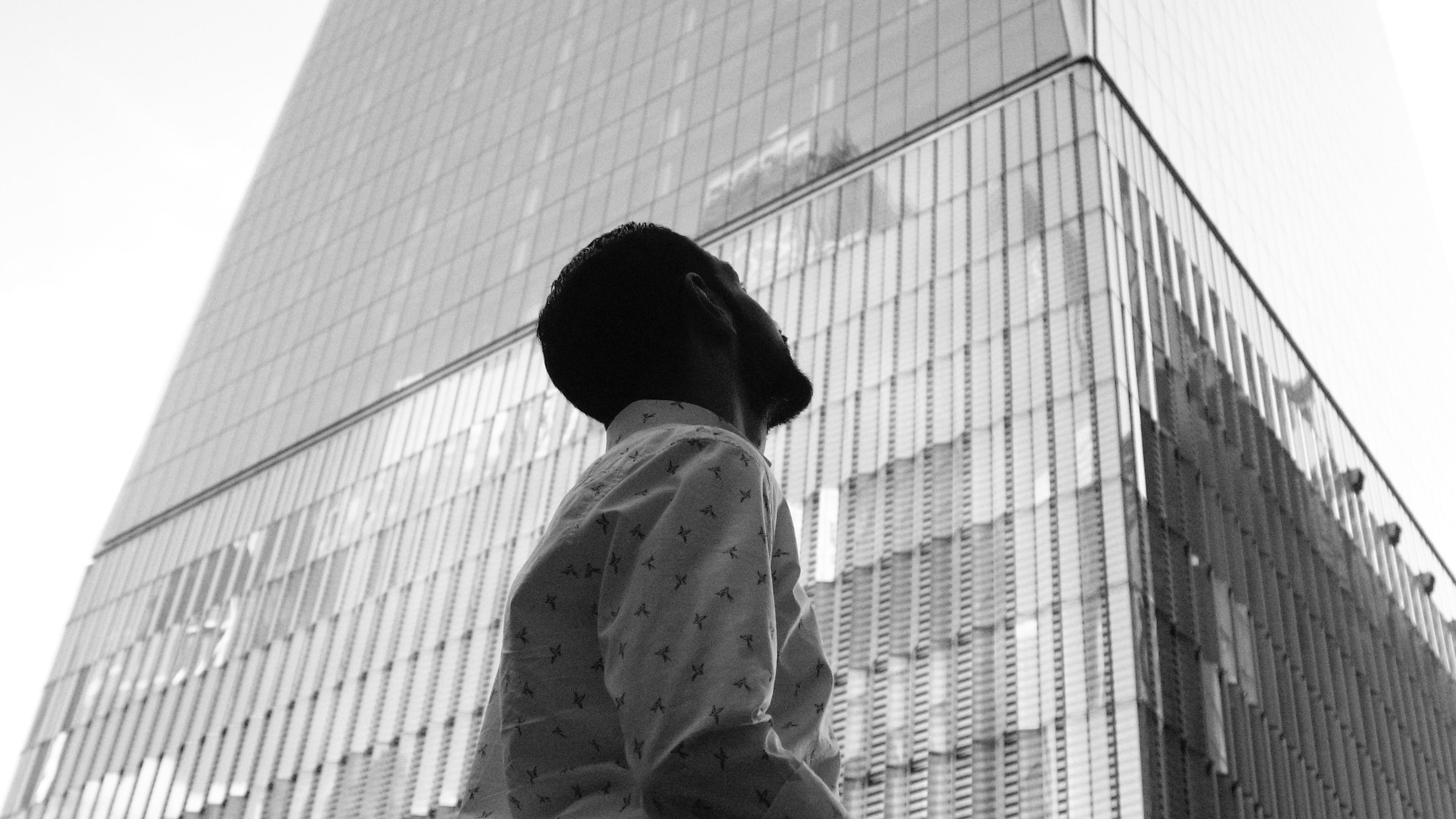 Black and white photo of a person looking up at a tall glass skyscraper with a suitcase in hand.
