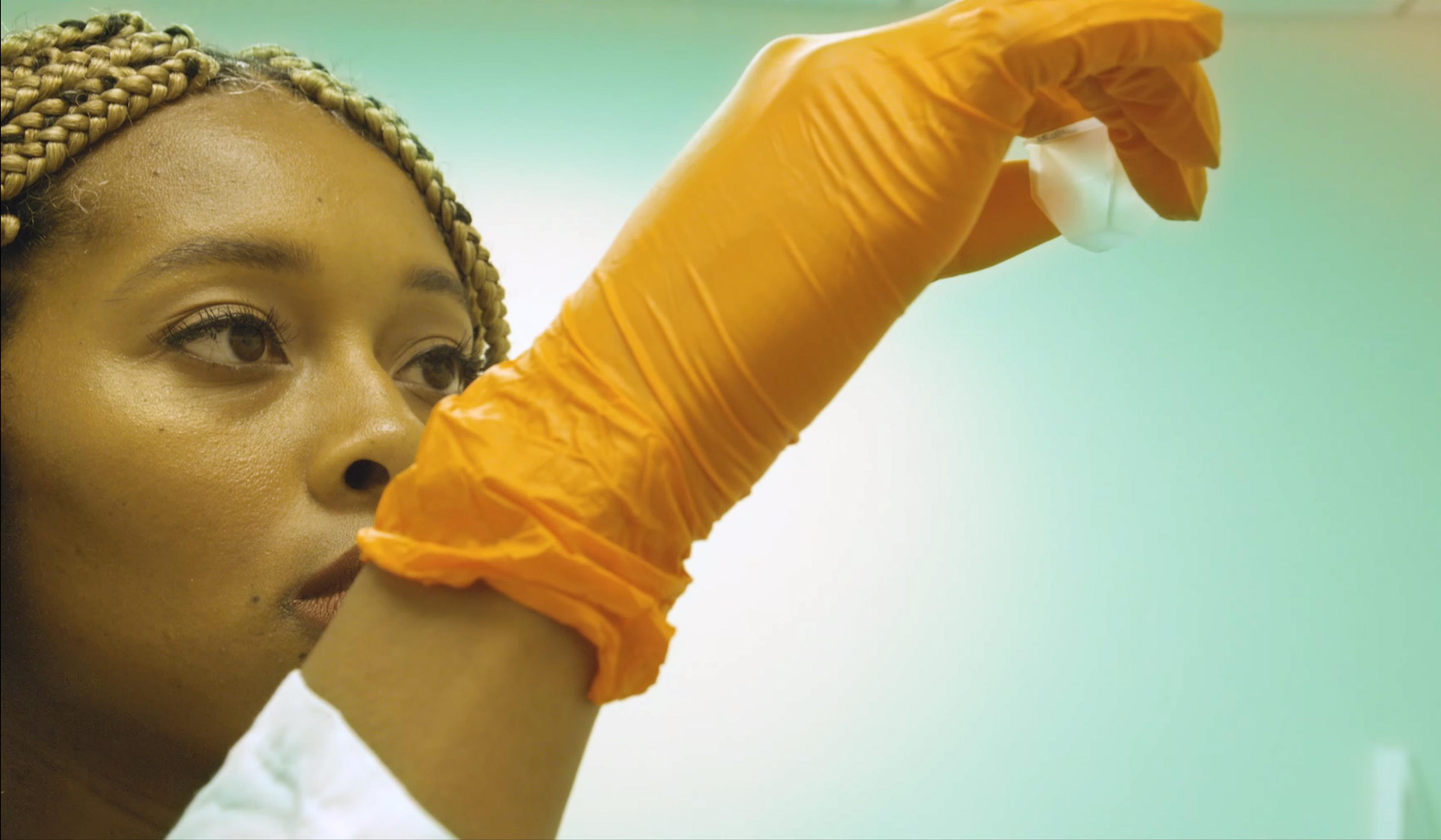 Photo of a scientist with braided hair examining a sample. She wears orange gloves and looks attentively at the object in a laboratory.