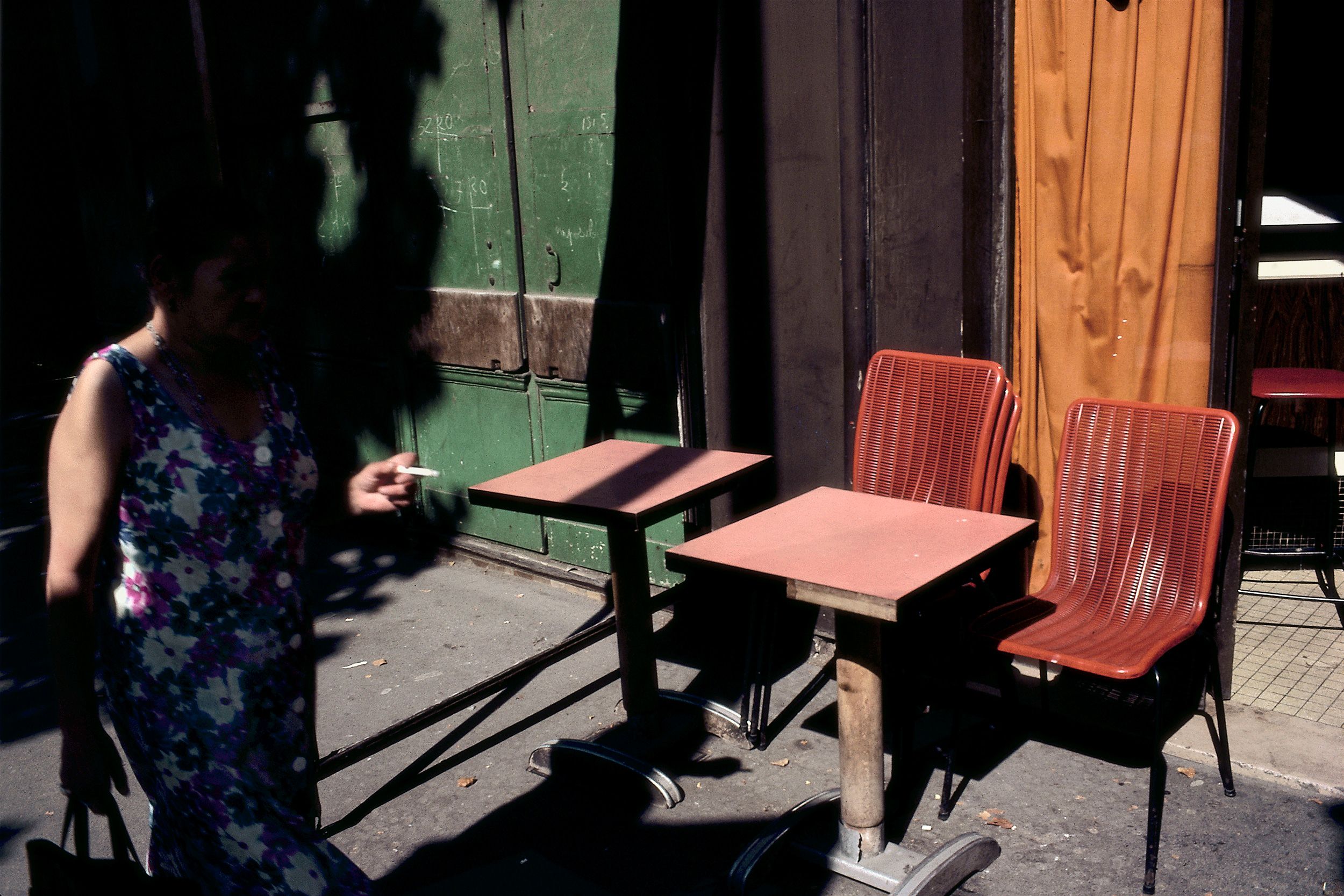 A person walking past outdoor café tables and chairs in shadowed sunlight.