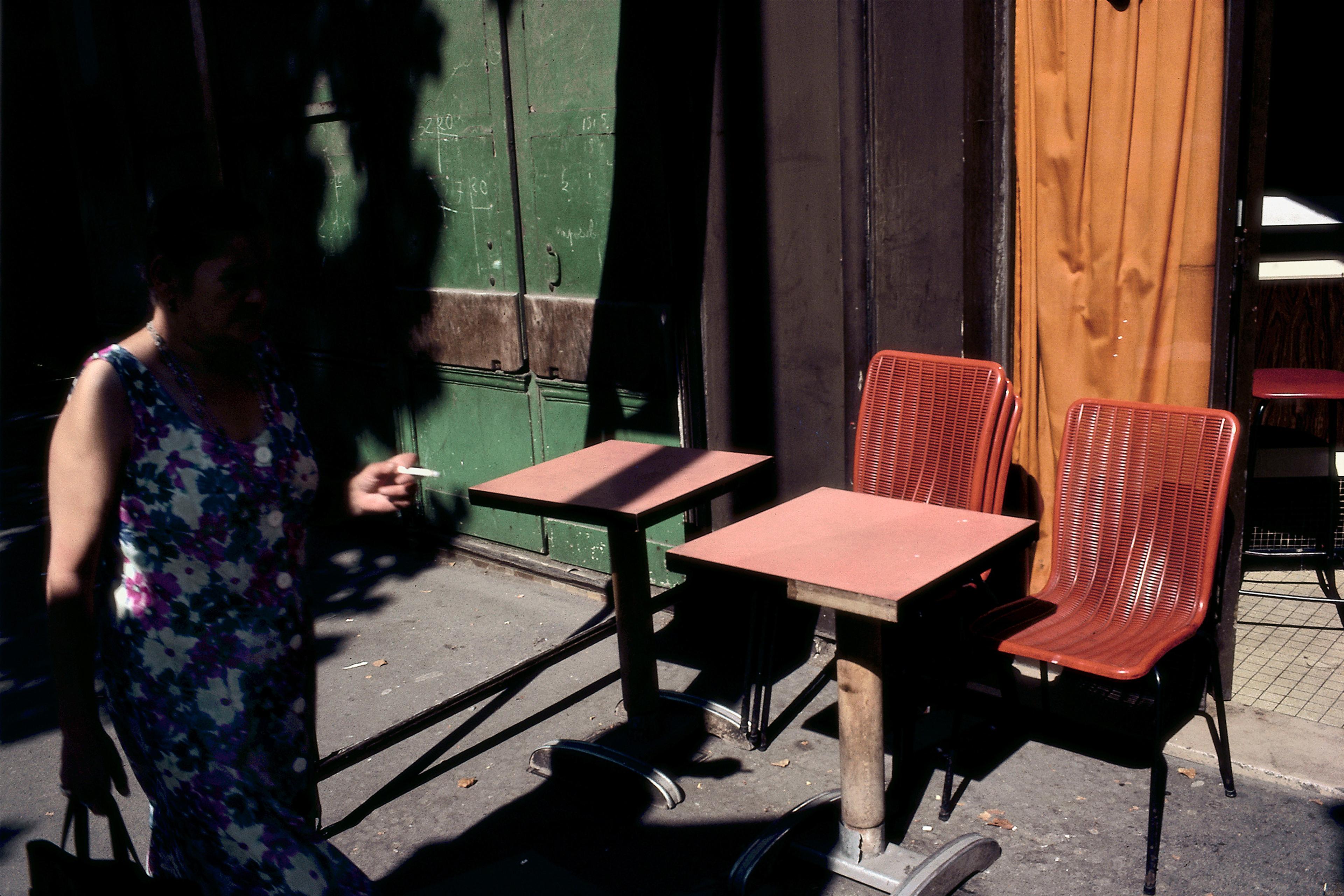 A person walking past outdoor café tables and chairs in shadowed sunlight.