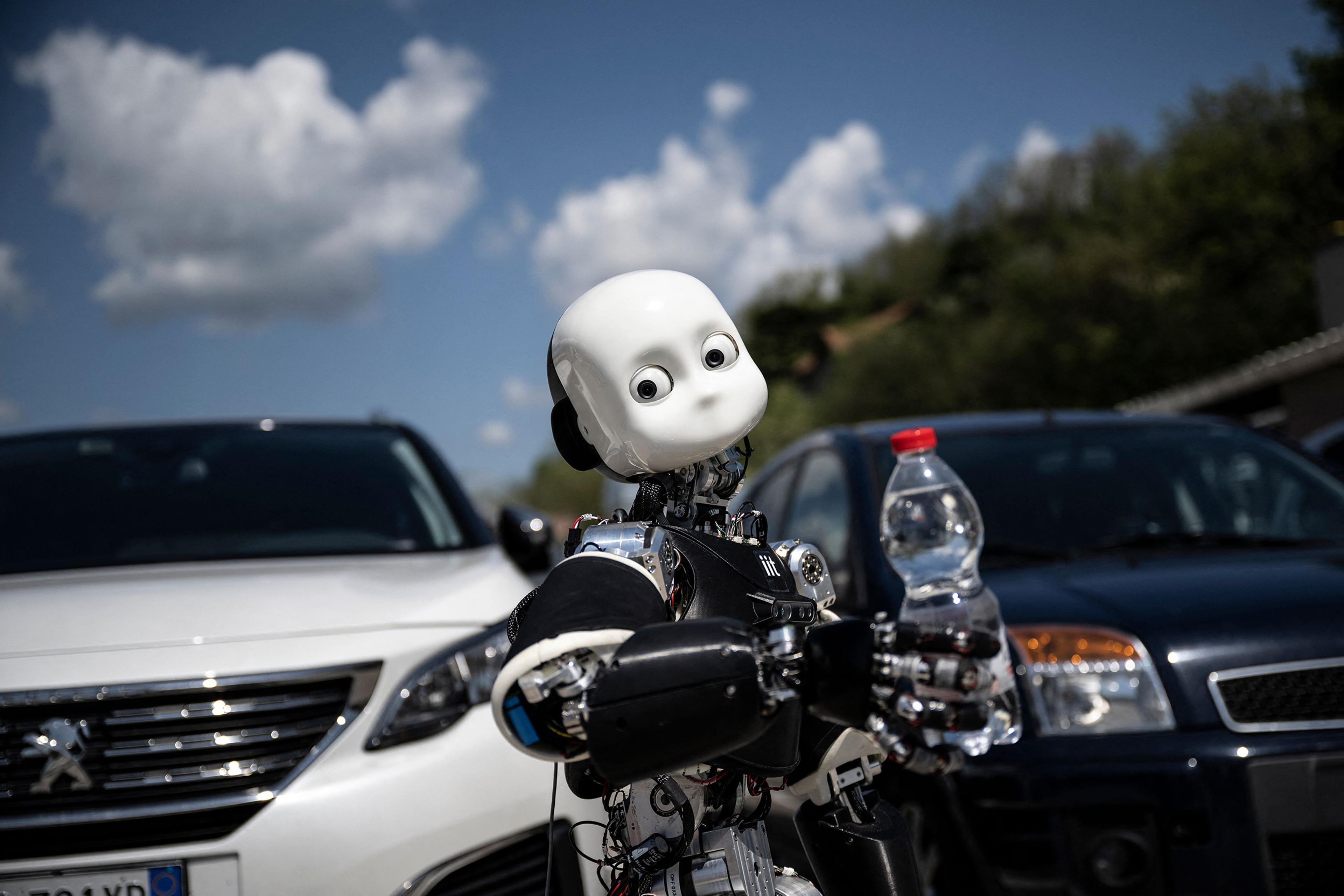 A humanoid robot holding a water bottle in front of parked cars under a partly cloudy sky.
