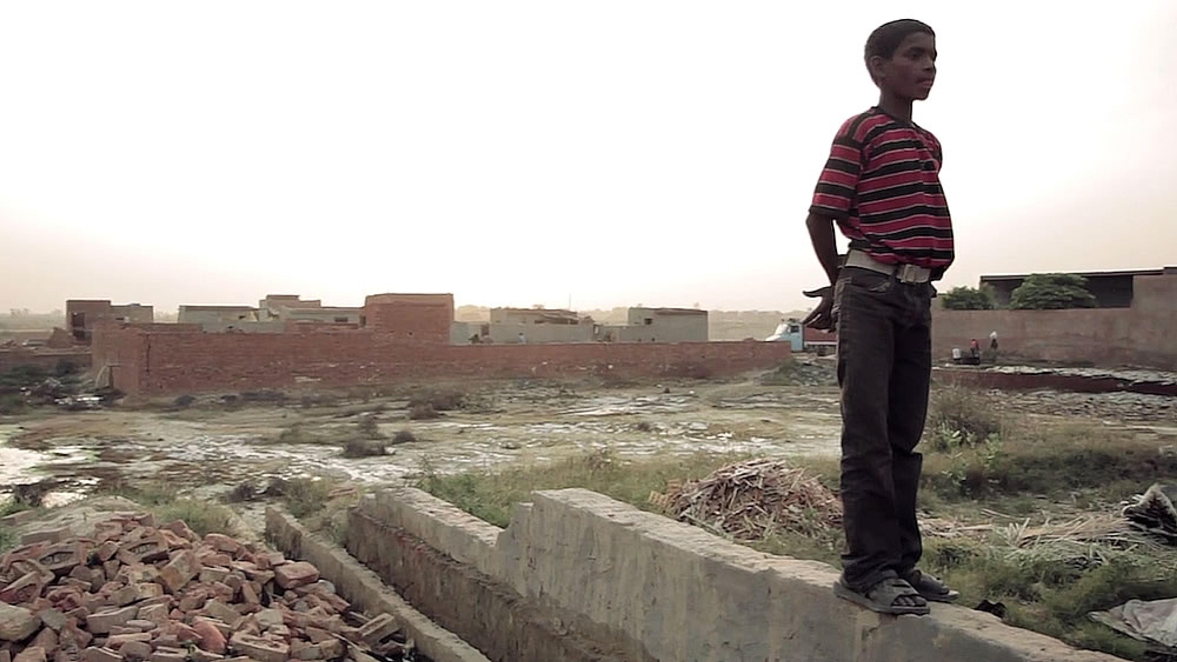 A boy in a red striped shirt and jeans standing on a wall, overlooking a field with buildings in the background at sunset.