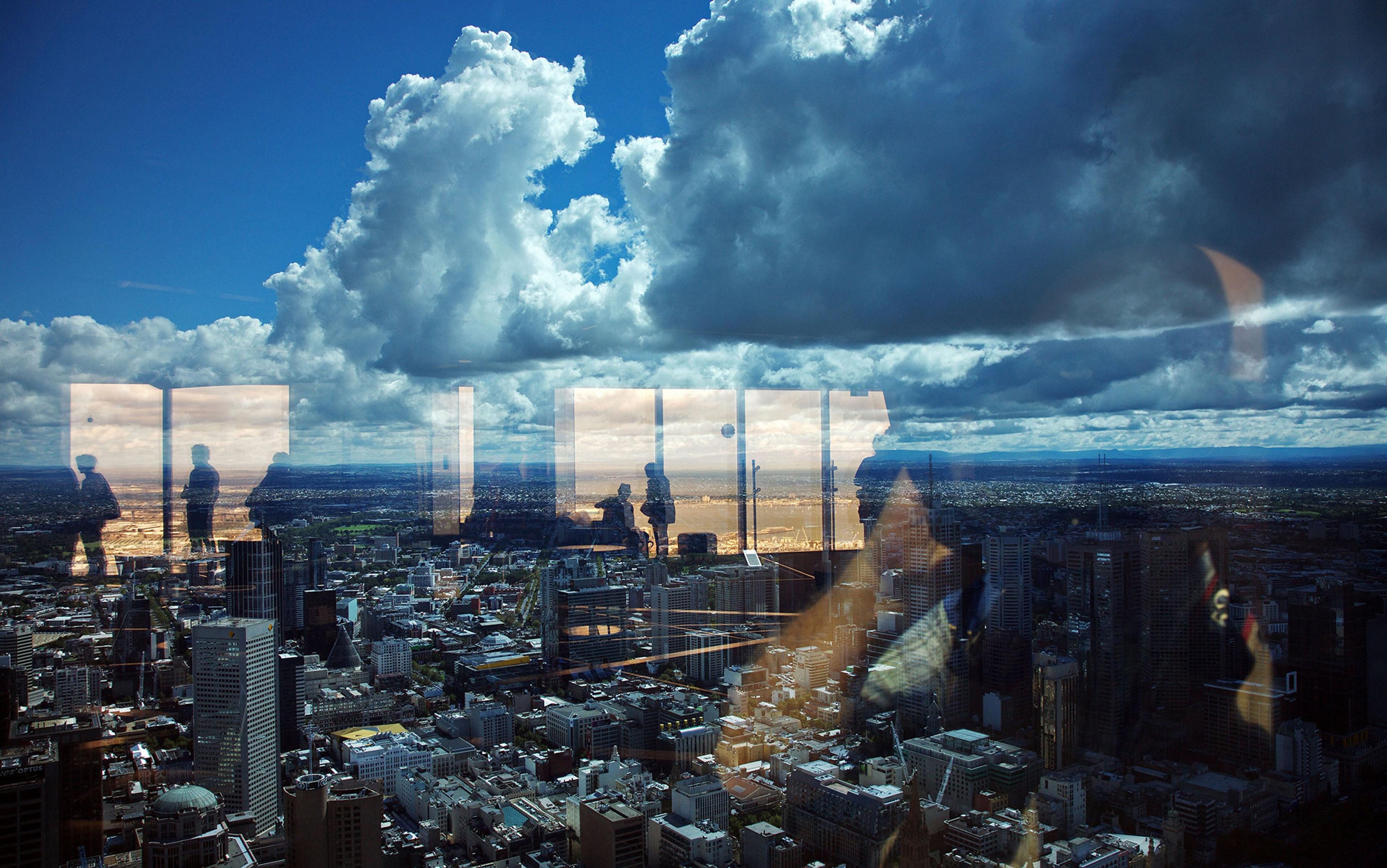 A cityscape view with reflections of people on windows and a dramatic cloudy sky in the background.