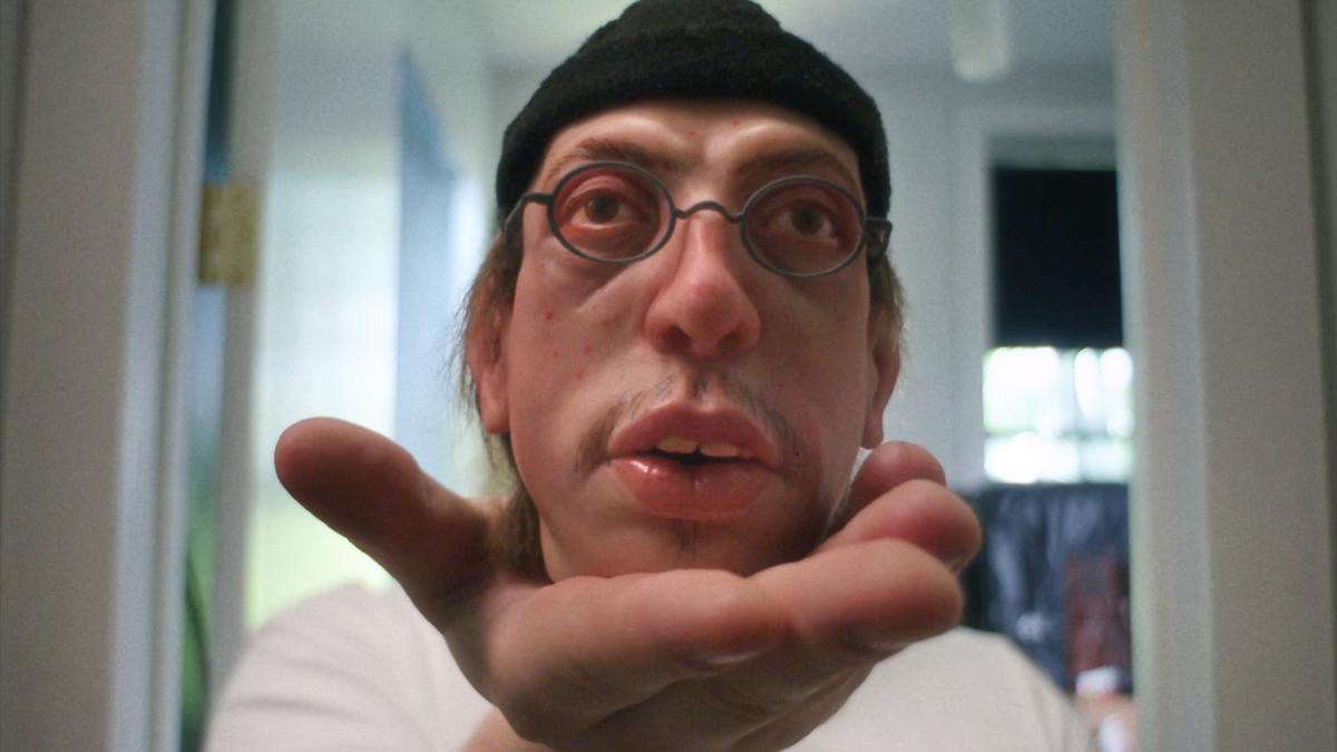 Surreal photo of a person wearing glasses and a hat with their face distorted, leaning towards a camera in a hallway.