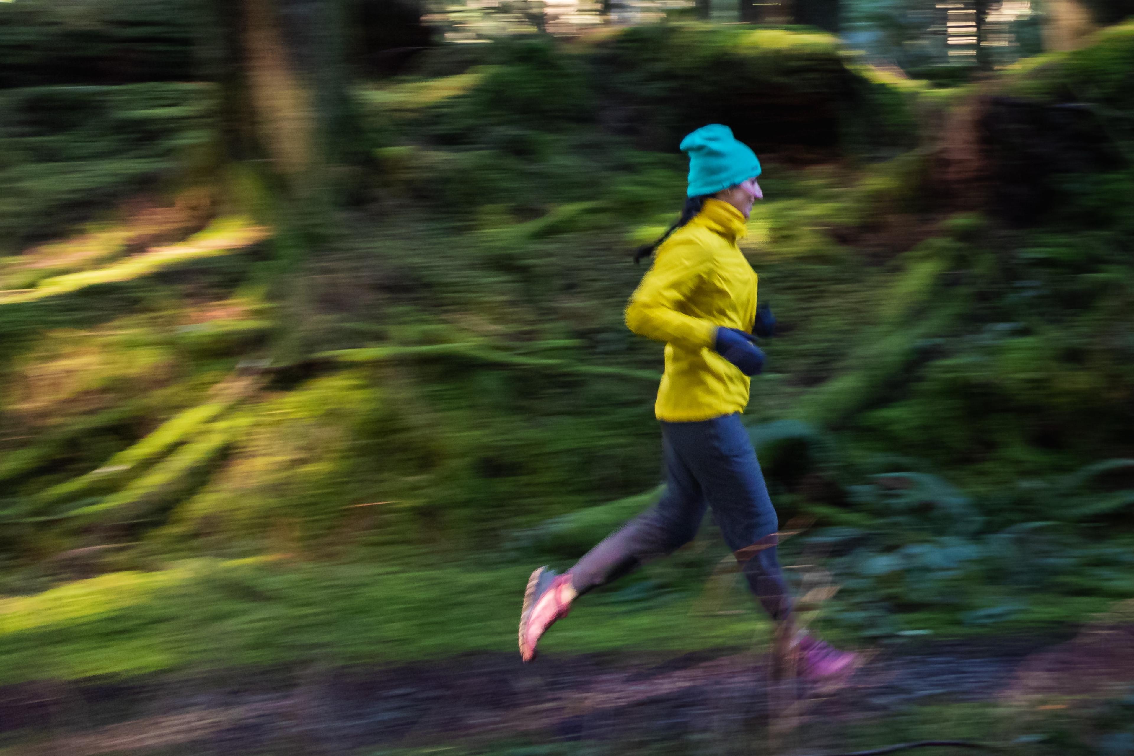 Photo of a person in a yellow jacket and turquoise hat running through a blurred forest path, conveying motion.