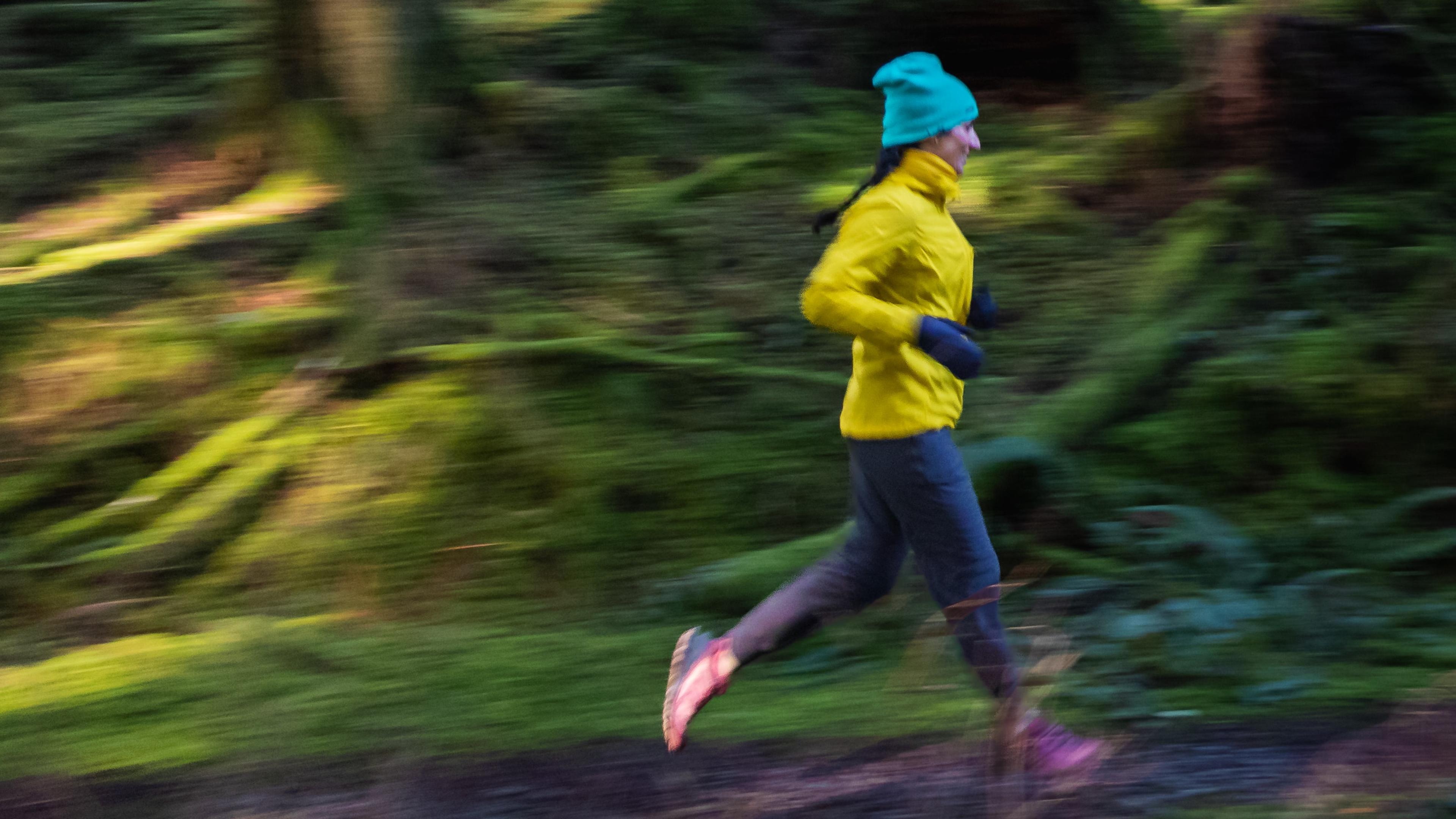 Photo of a person in a yellow jacket and turquoise hat running through a blurred forest path, conveying motion.