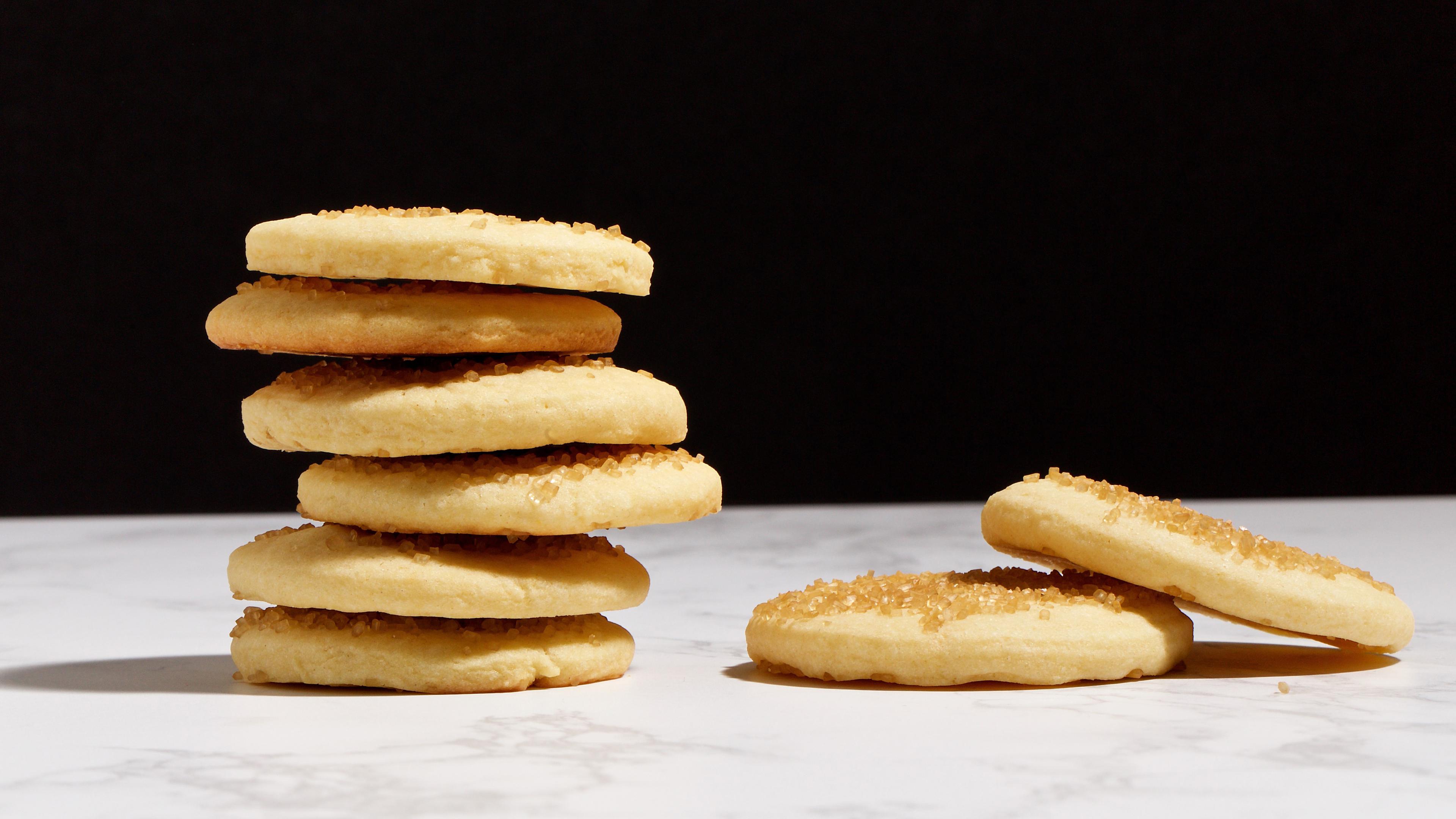 Photo of a stack of six sugar cookies on a marble surface with two cookies lying flat beside them against a dark background.