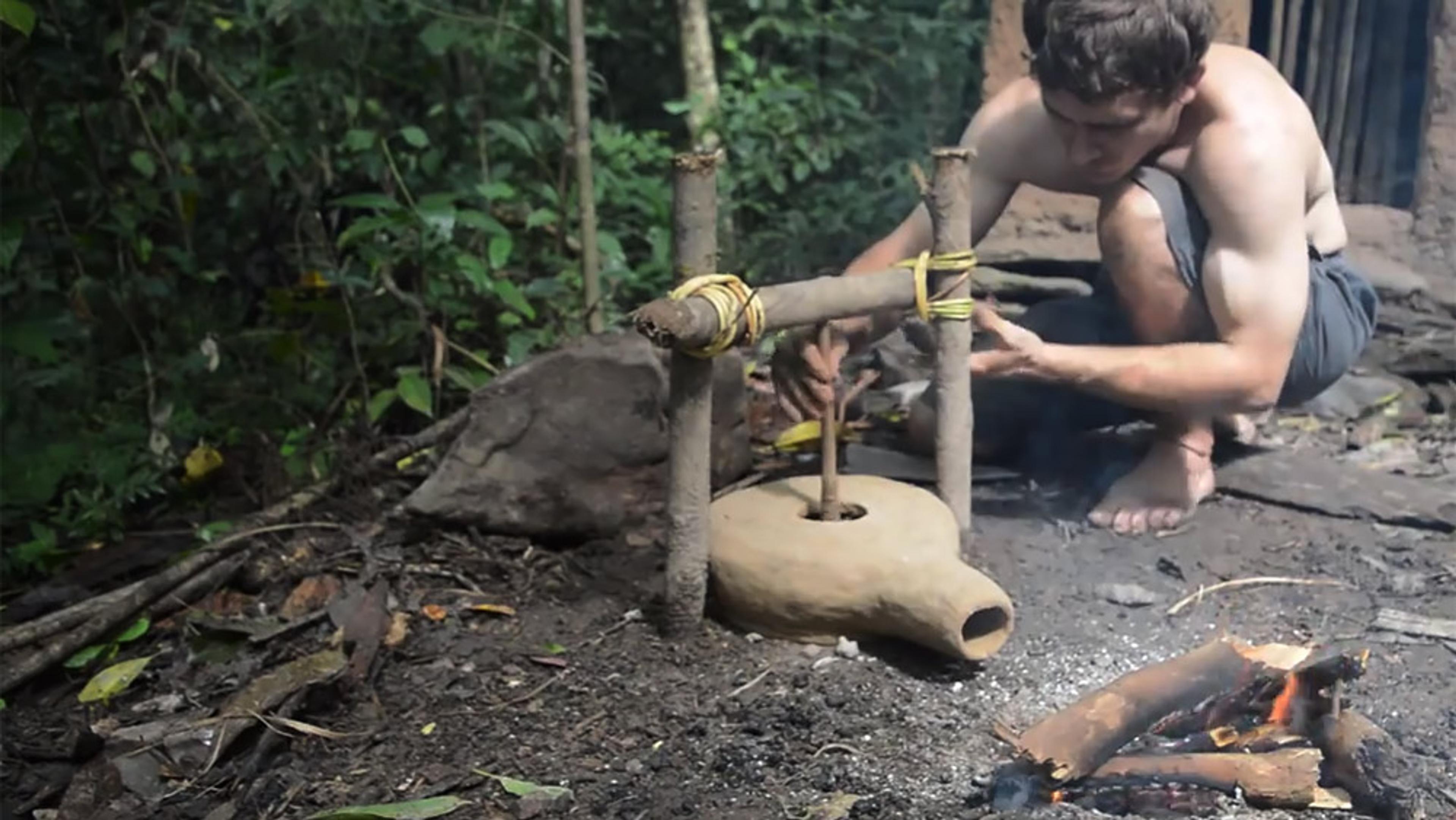 A man in shorts using a handmade air-blower device to stoke a fire in a wooded outdoor setting.