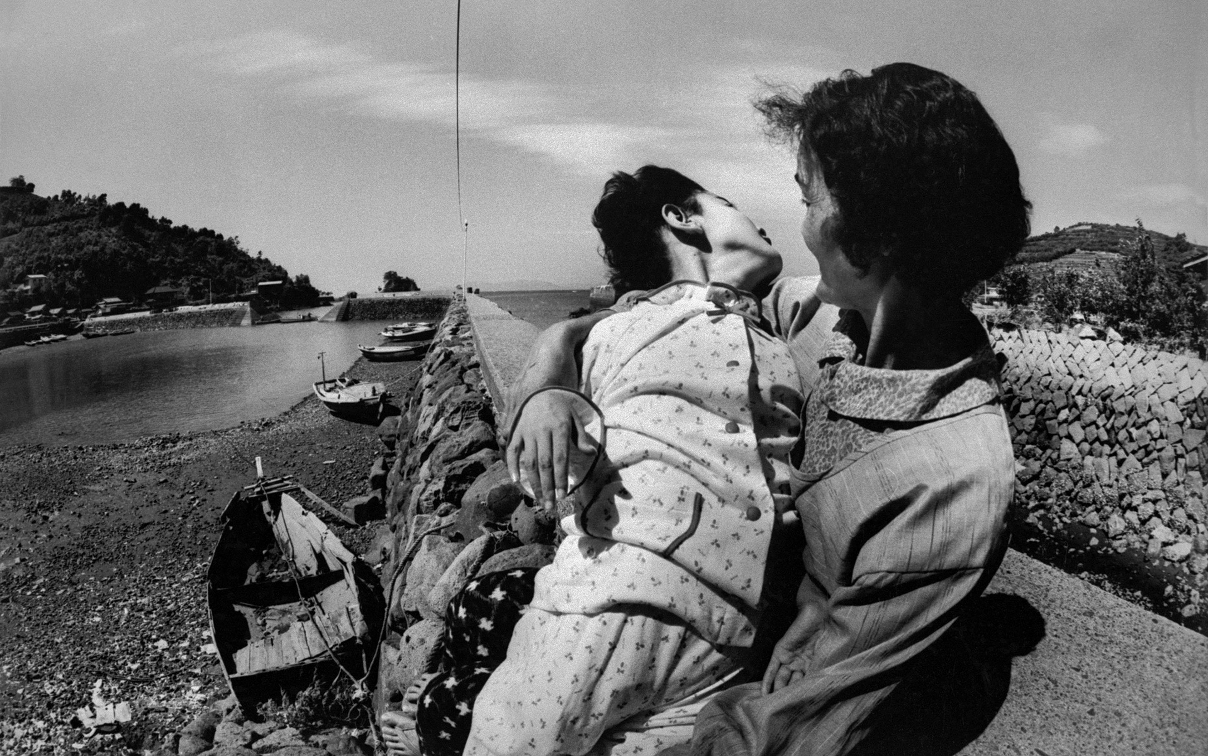 Black-and-white photo of a woman holding her child on a stone pier, with small wooden boats and a shoreline in the background.