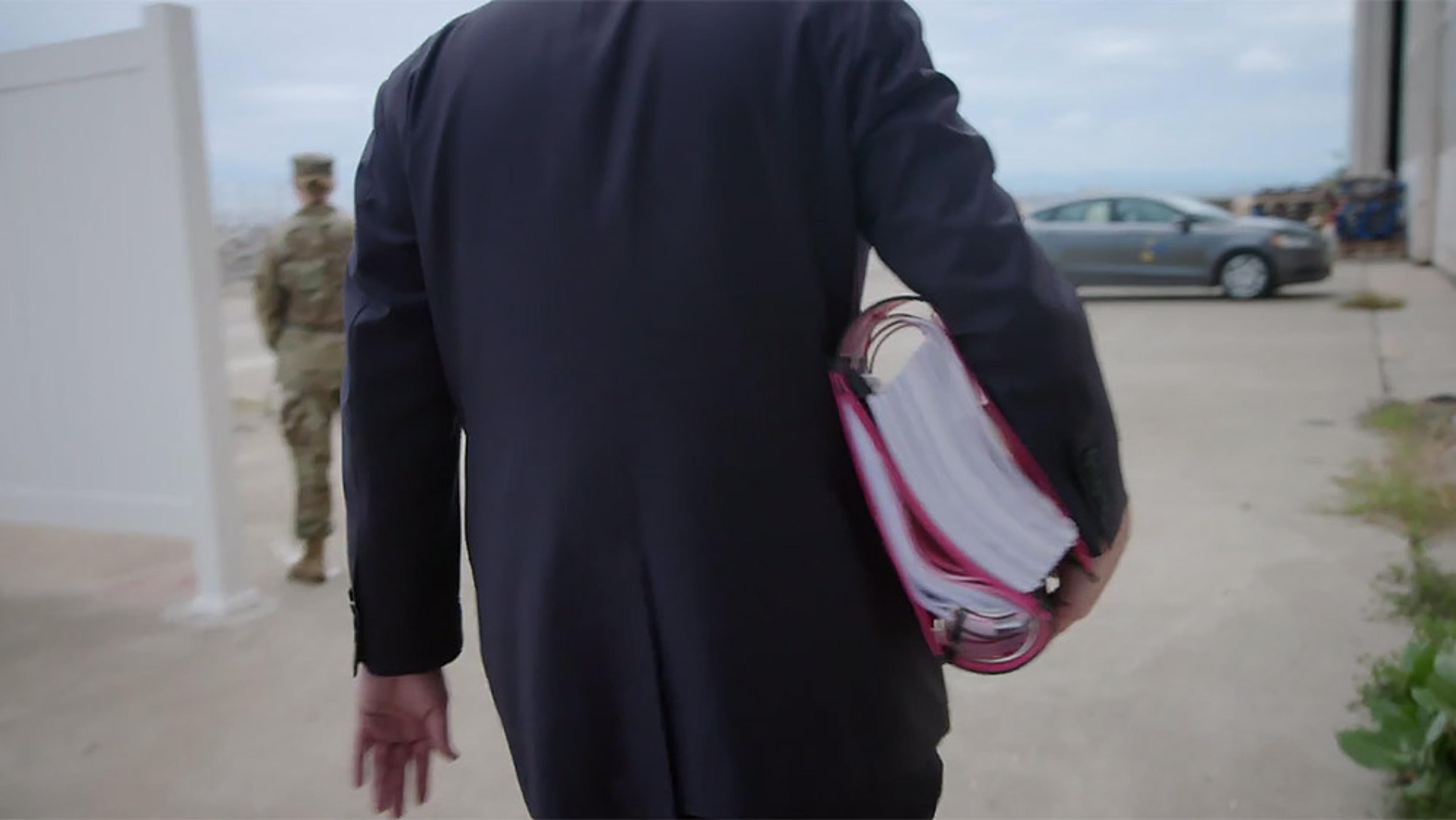 A person in a suit carrying a large binder. A person in military uniform stands nearby in an outdoor setting with a parked car.