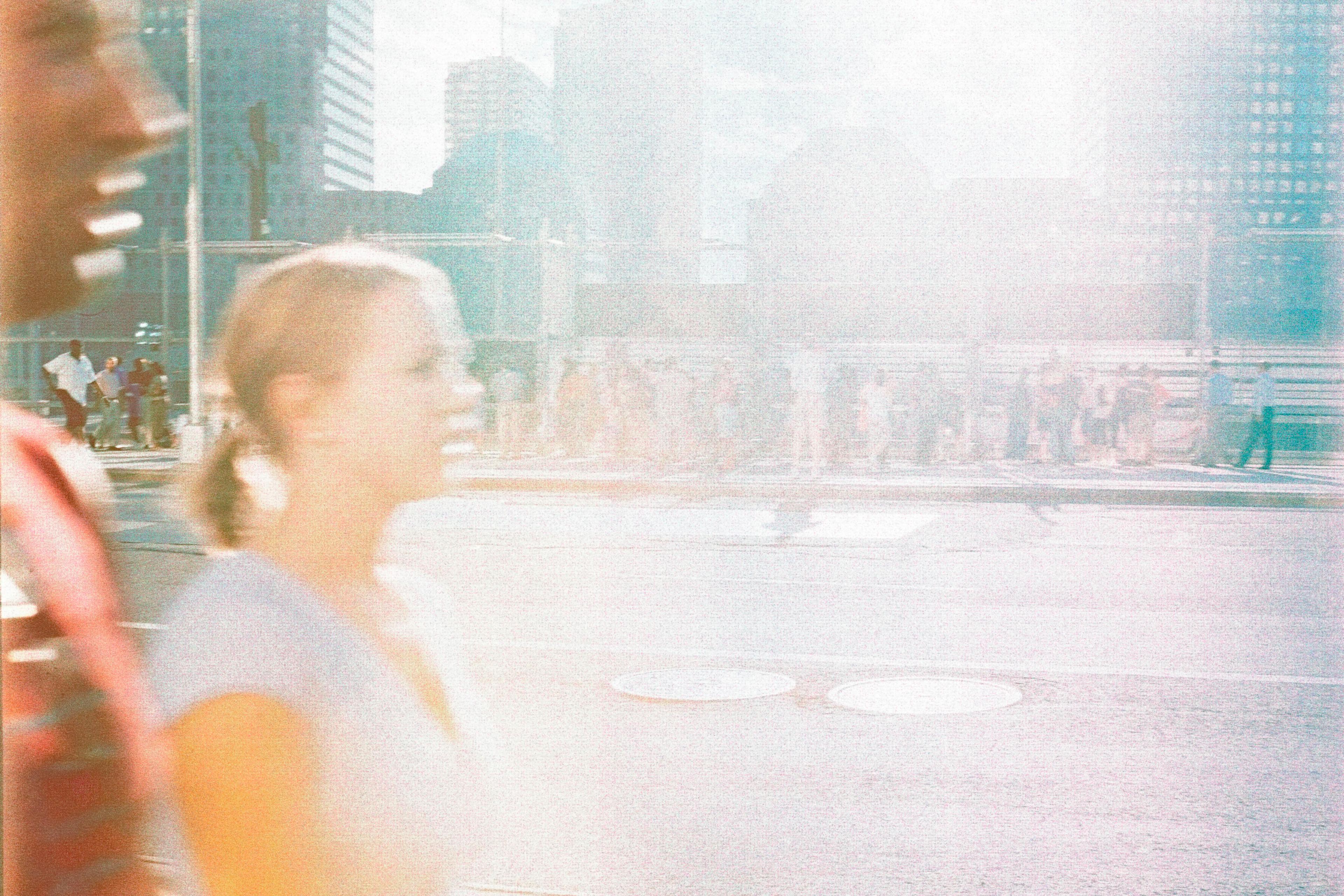 Photo of a city street with two blurry people walking in foreground bright light flaring over the scene in the afternoon.